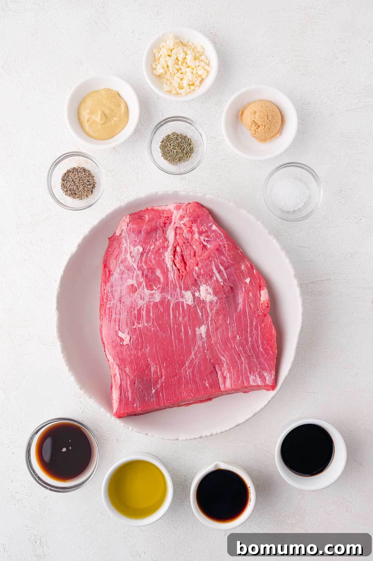 Raw flank steak on a plate surrounded by small bowls containing garlic, mustard, brown sugar, salt, pepper, dried herbs, soy sauce, olive oil, and Worcestershire sauce.