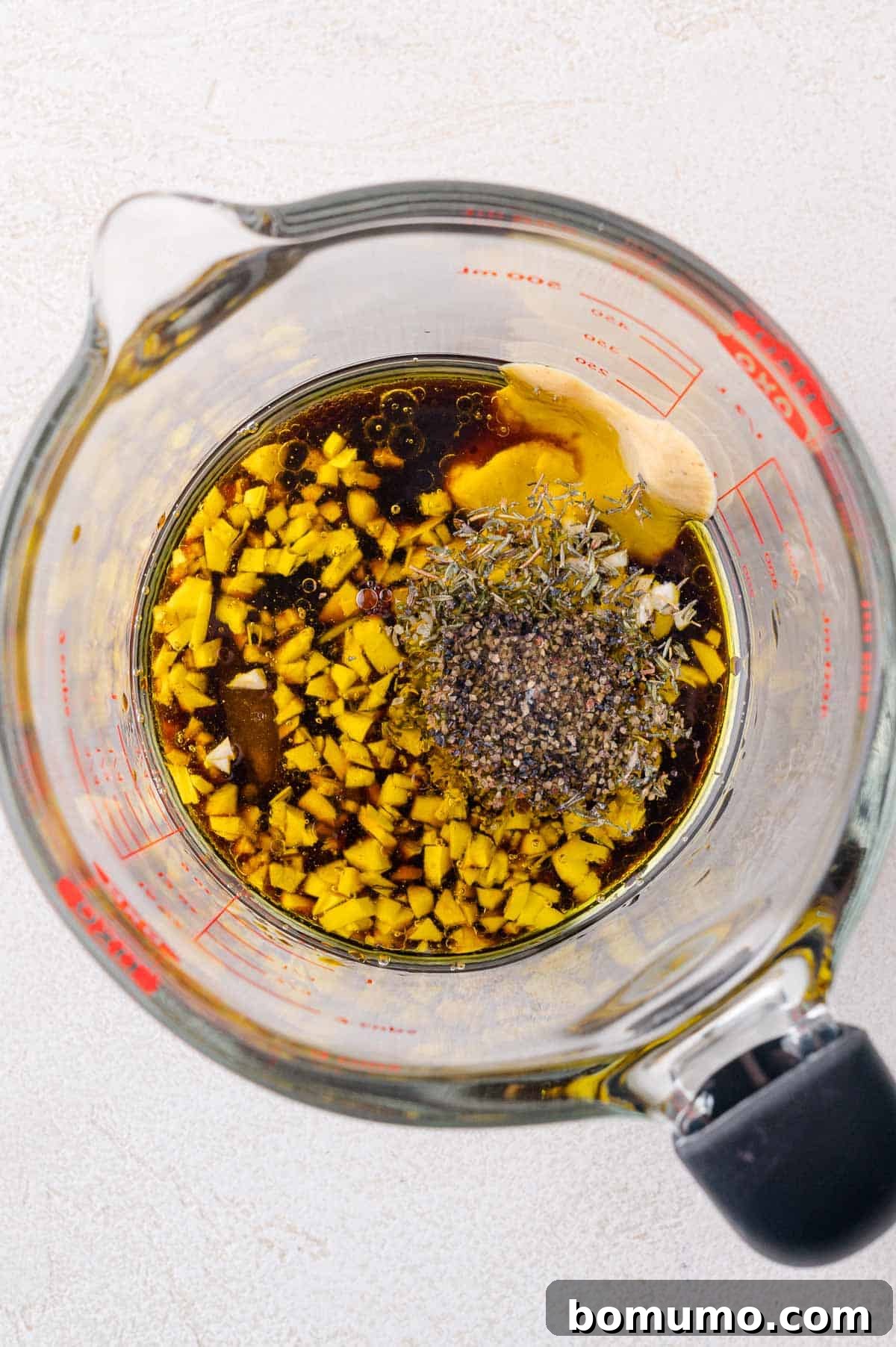 A glass measuring cup filled with chopped garlic, black pepper, dried herbs, soy sauce, oil, and vinegar for making a marinade.