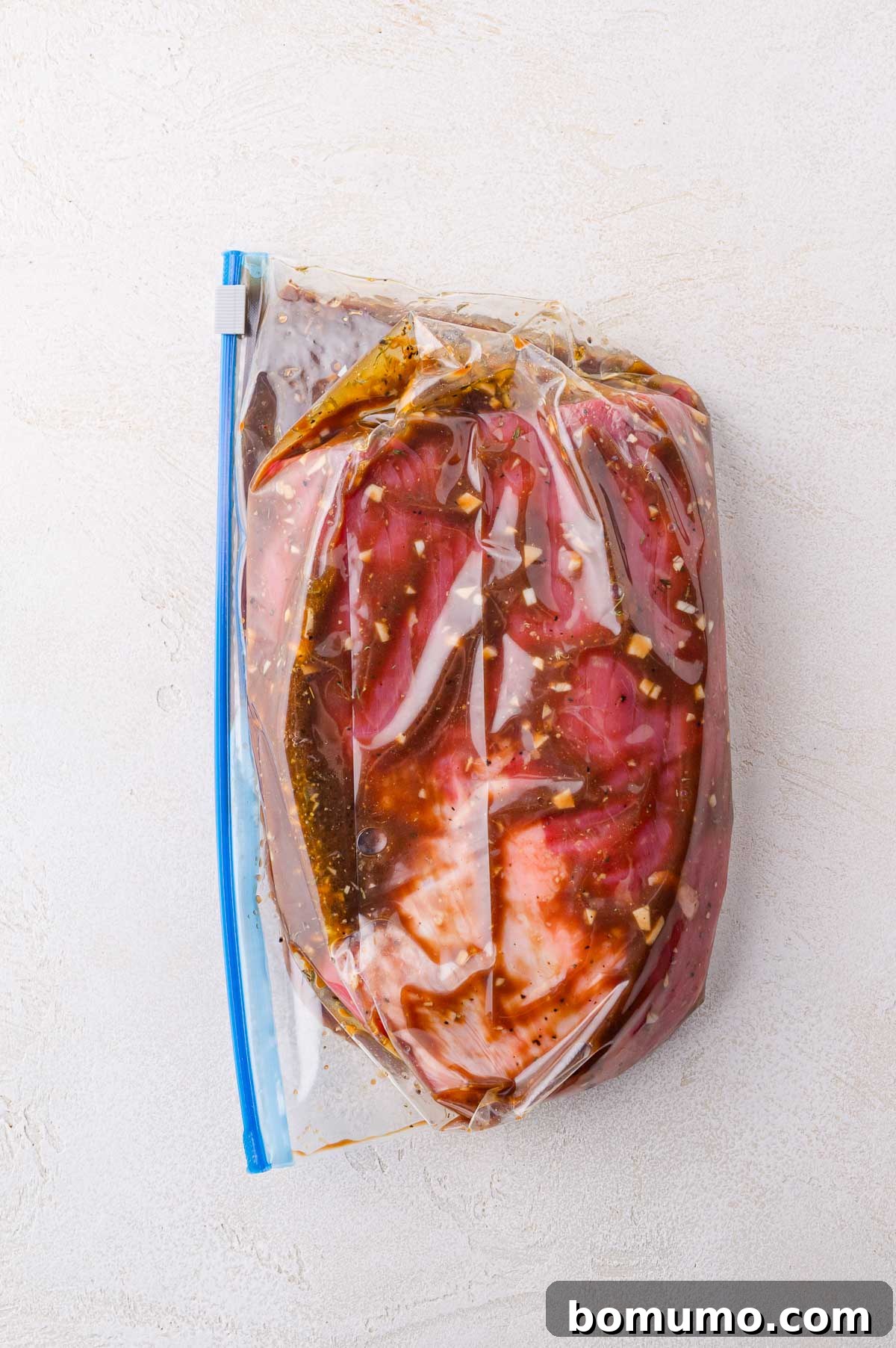 A piece of raw meat marinating in a clear plastic zip-top bag with visible herbs and sauce, placed on a light-colored surface.