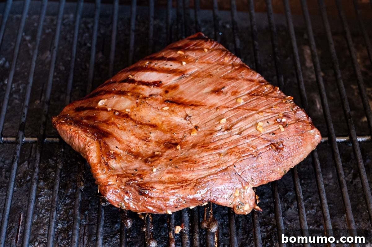 A piece of grilled flank steak with sear marks is cooking on a barbecue grill.