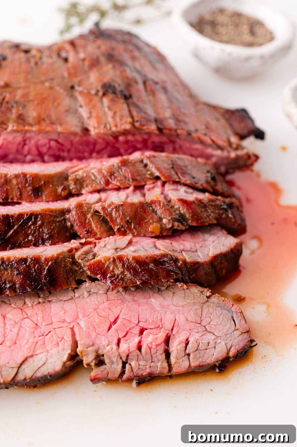 Sliced medium-rare grilled steak on a white surface, with juices visible and a bowl of black pepper in the background.