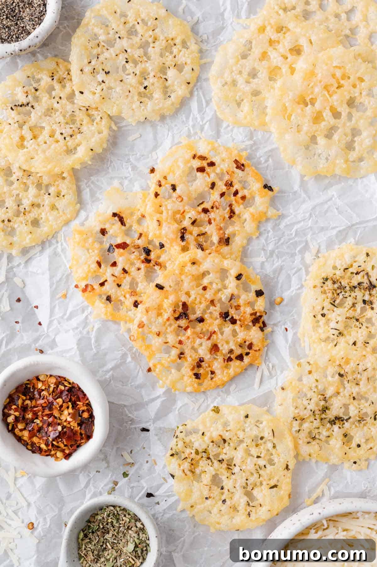 Crispy cheese rounds rest on crumpled parchment paper, surrounded by small bowls containing chili flakes, dried herbs, and shredded cheese, ready to be seasoned.