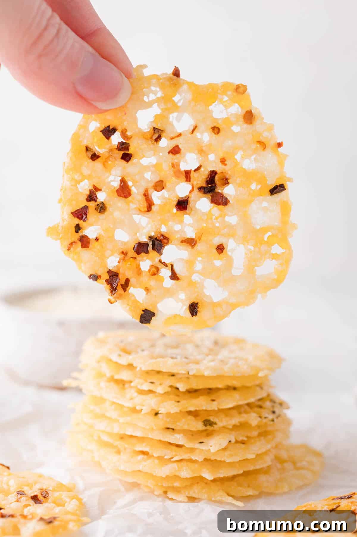A hand holds up a thin, round cheese crisp topped with red pepper flakes above a stack of similar crisps on a white surface, highlighting its delicate texture.