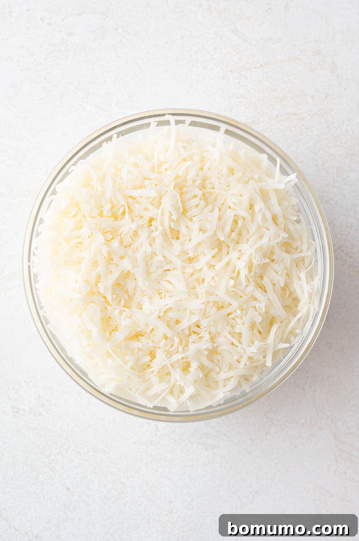 A clear glass bowl filled with freshly shredded Parmesan cheese sits on a clean white surface, showcasing the quality of the ingredient.