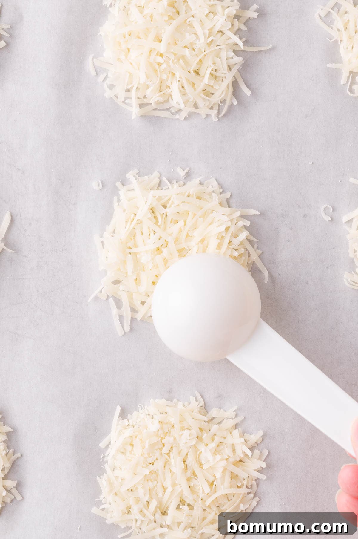 A white measuring spoon holds shredded cheese, meticulously forming small, even piles on parchment paper, demonstrating precise portioning.