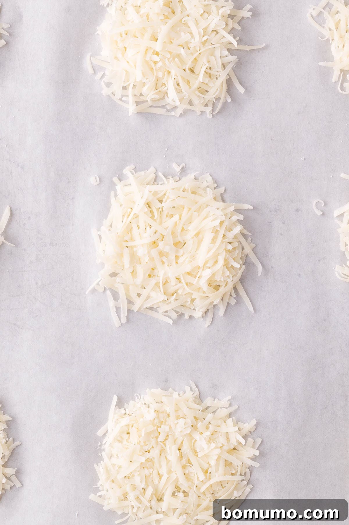 Shredded Parmesan cheese is arranged in small, evenly spaced mounds on a sheet of parchment paper, ready for baking.