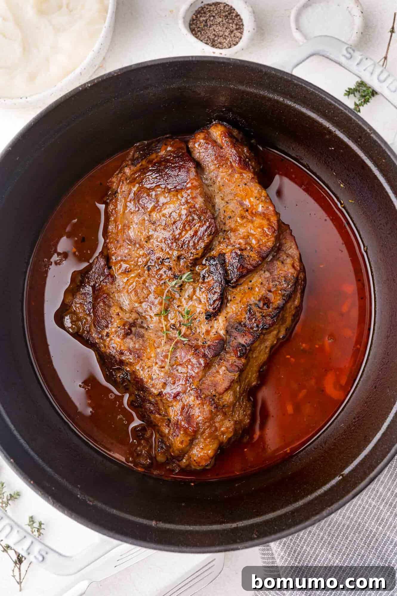 Roasted meat in a black pot with sauce, garnished with thyme, placed on a light surface with a napkin and bowls nearby.
