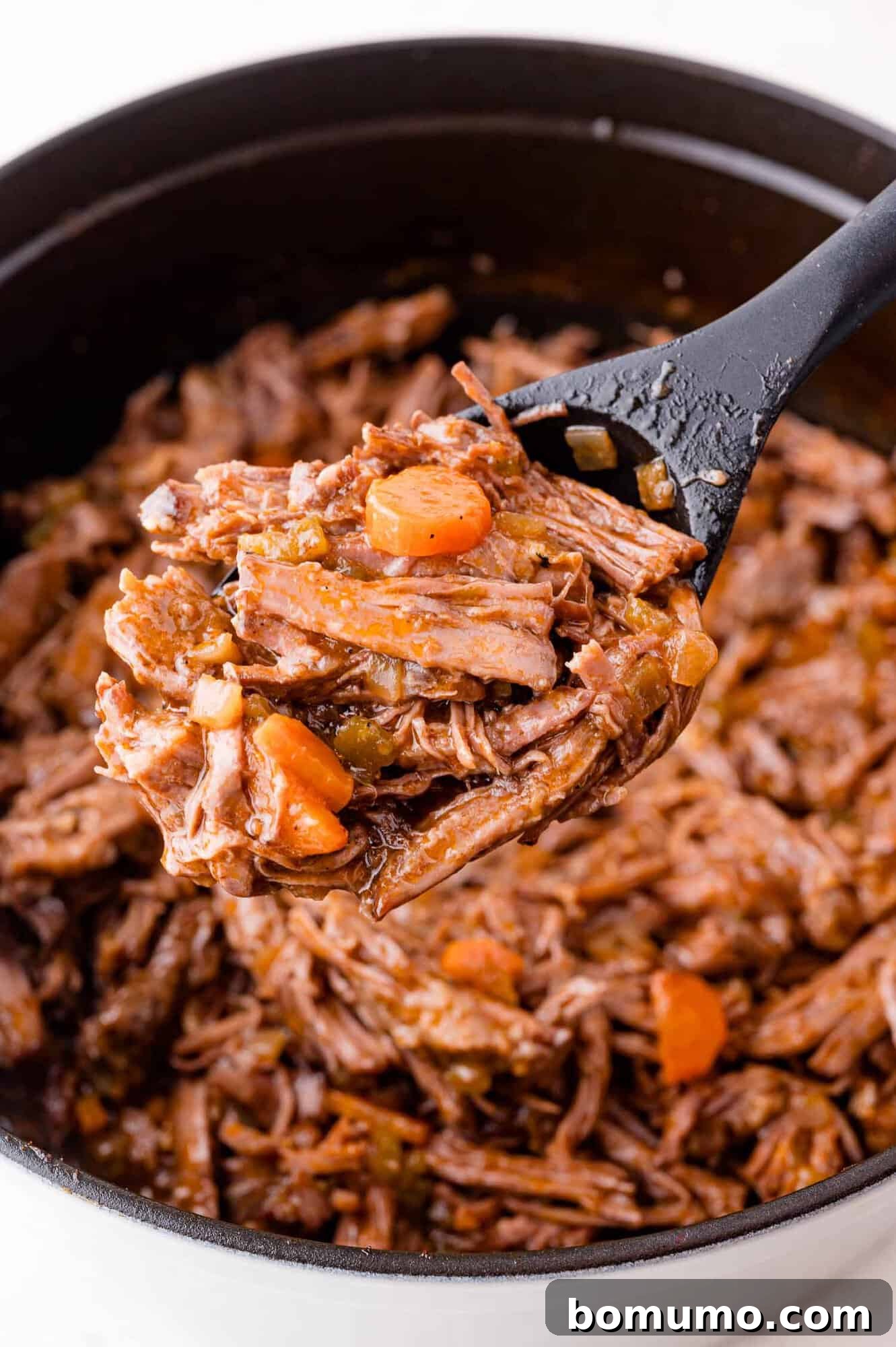 Close-up of shredded braised beef with carrots and onions in a black pot, being scooped with a black utensil.