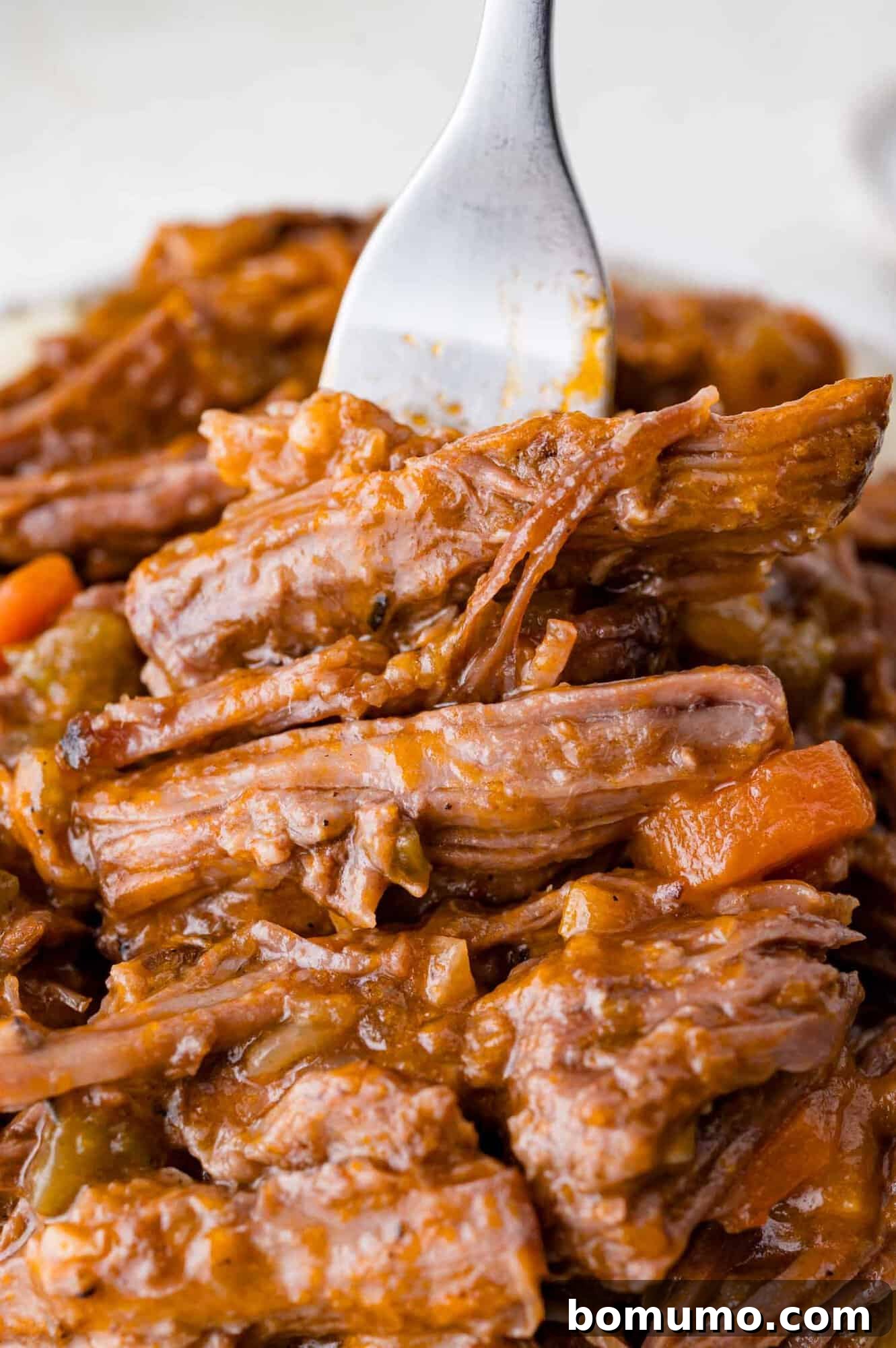 A fork holds a portion of shredded braised beef covered in gravy, with visible pieces of carrot and onion.