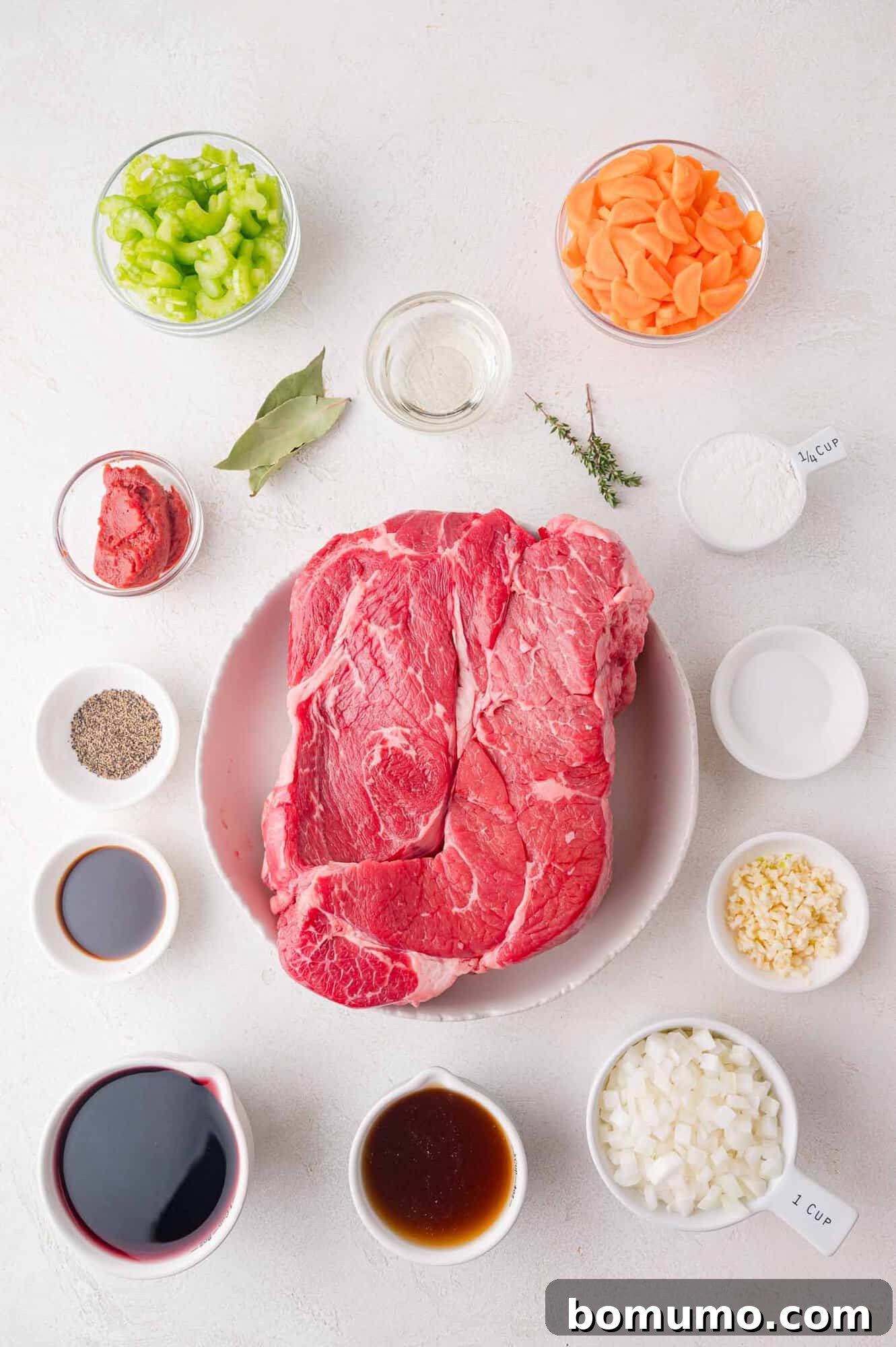 Raw beef on a plate surrounded by ingredients like chopped celery, carrots, onions, garlic, tomato paste, bay leaf, thyme, red wine, broth, and seasoning in bowls on a white surface.