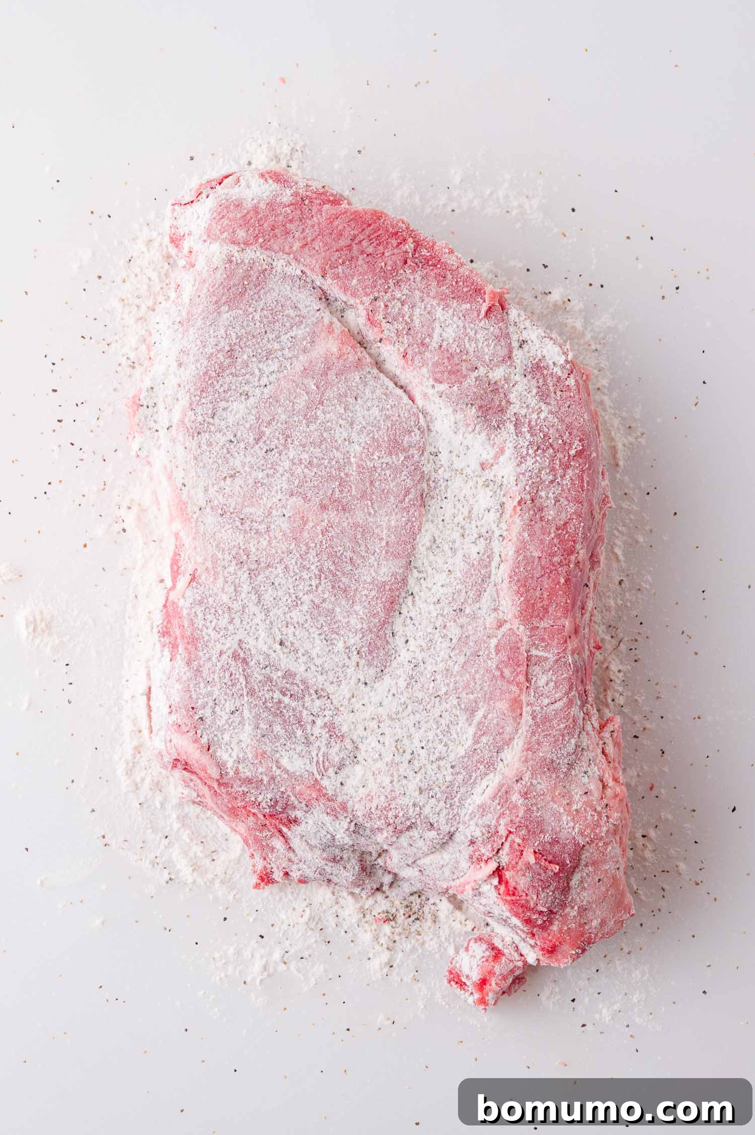 A raw piece of meat coated with white flour and black pepper, placed on a white surface.