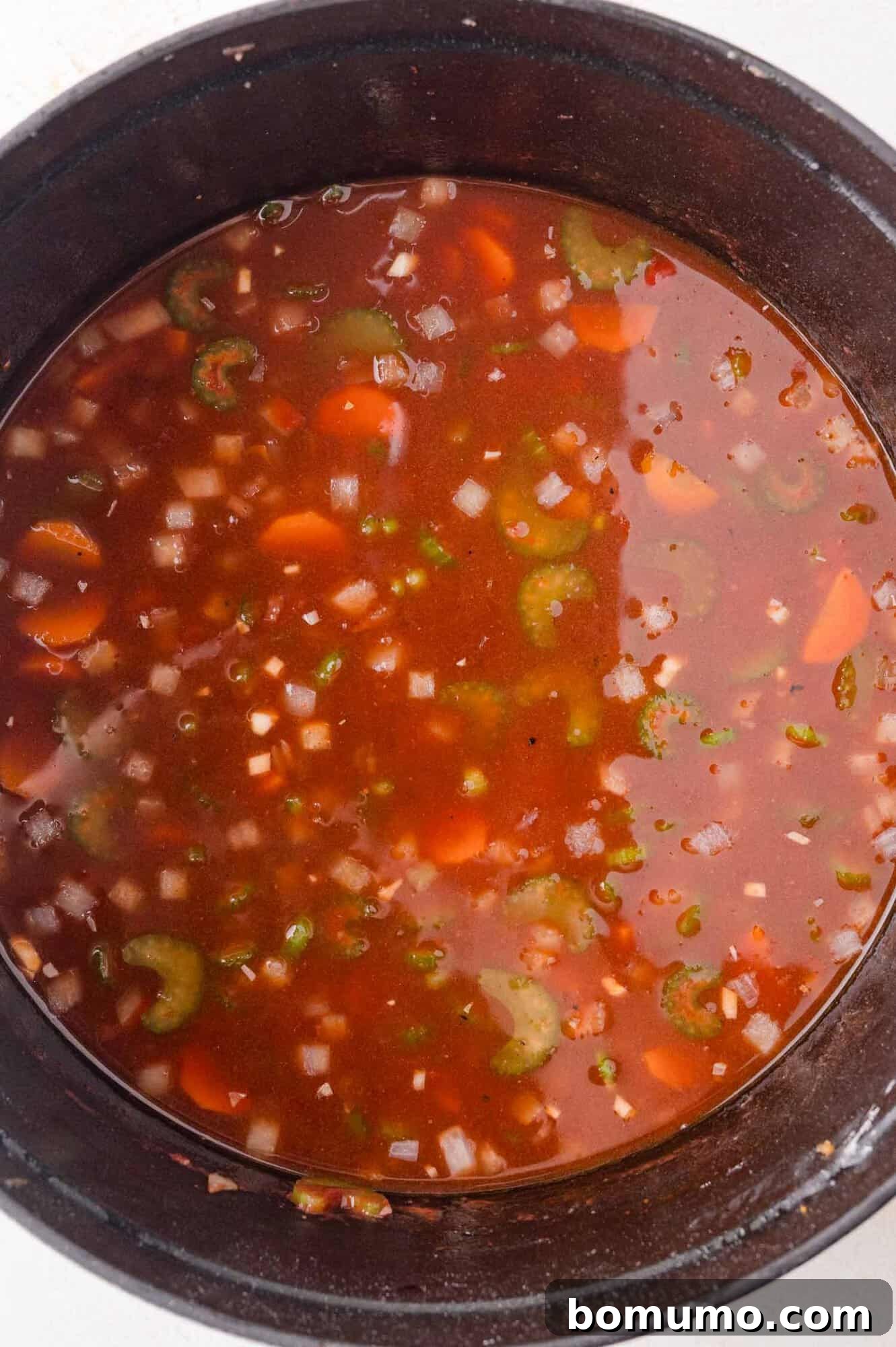 A pot containing visible pieces of carrots, celery, and onions in a reddish broth.