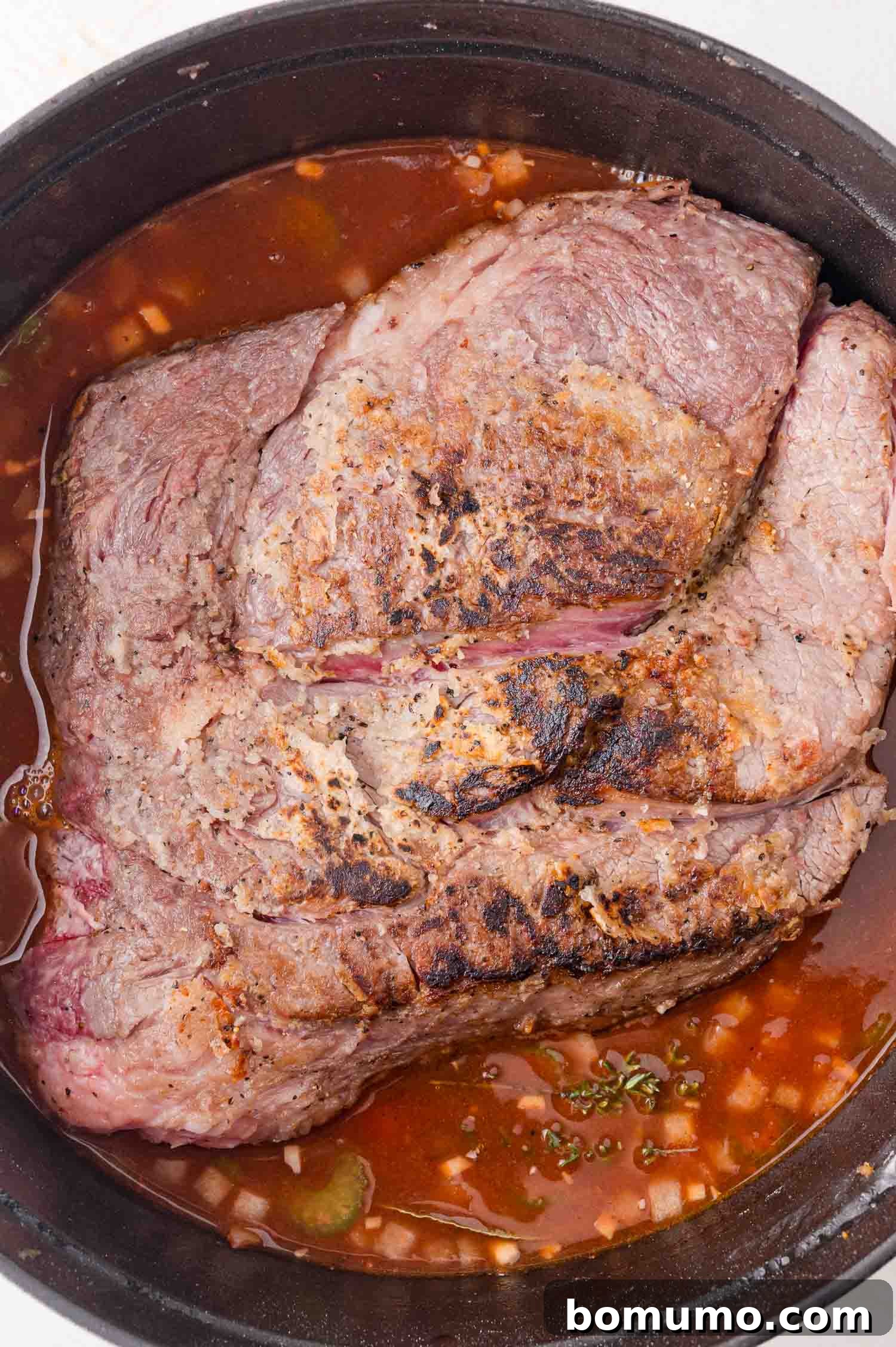 Browned beef roast in a black pot with sauce and diced onions.