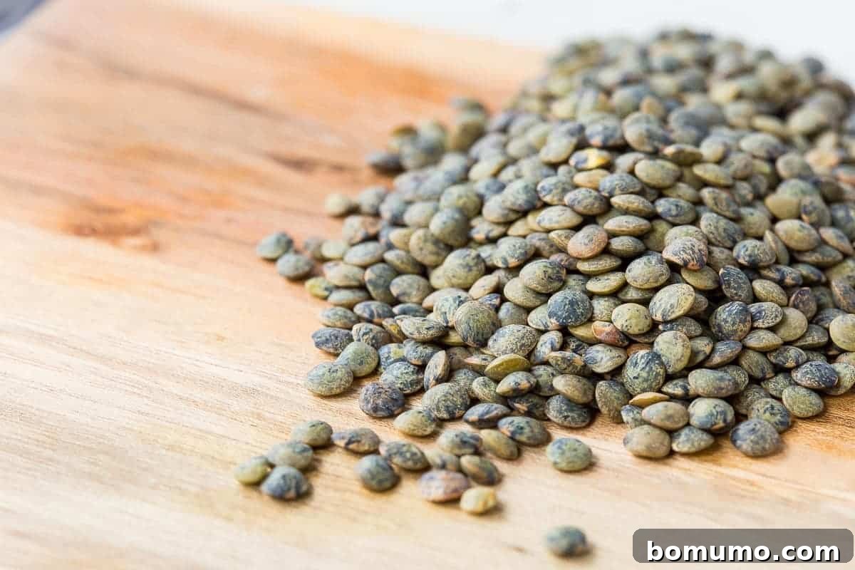 A scattering of beautiful, uncooked French green lentils spread across a rustic wooden cutting board, showcasing their distinct color and shape.