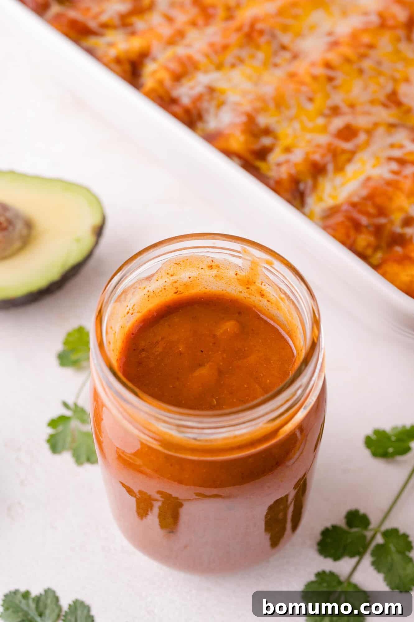 A jar of rich, homemade enchilada sauce on a rustic wooden table, with a halved avocado and fresh cilantro leaves in the foreground. Partially visible enchiladas hint at delicious possibilities.