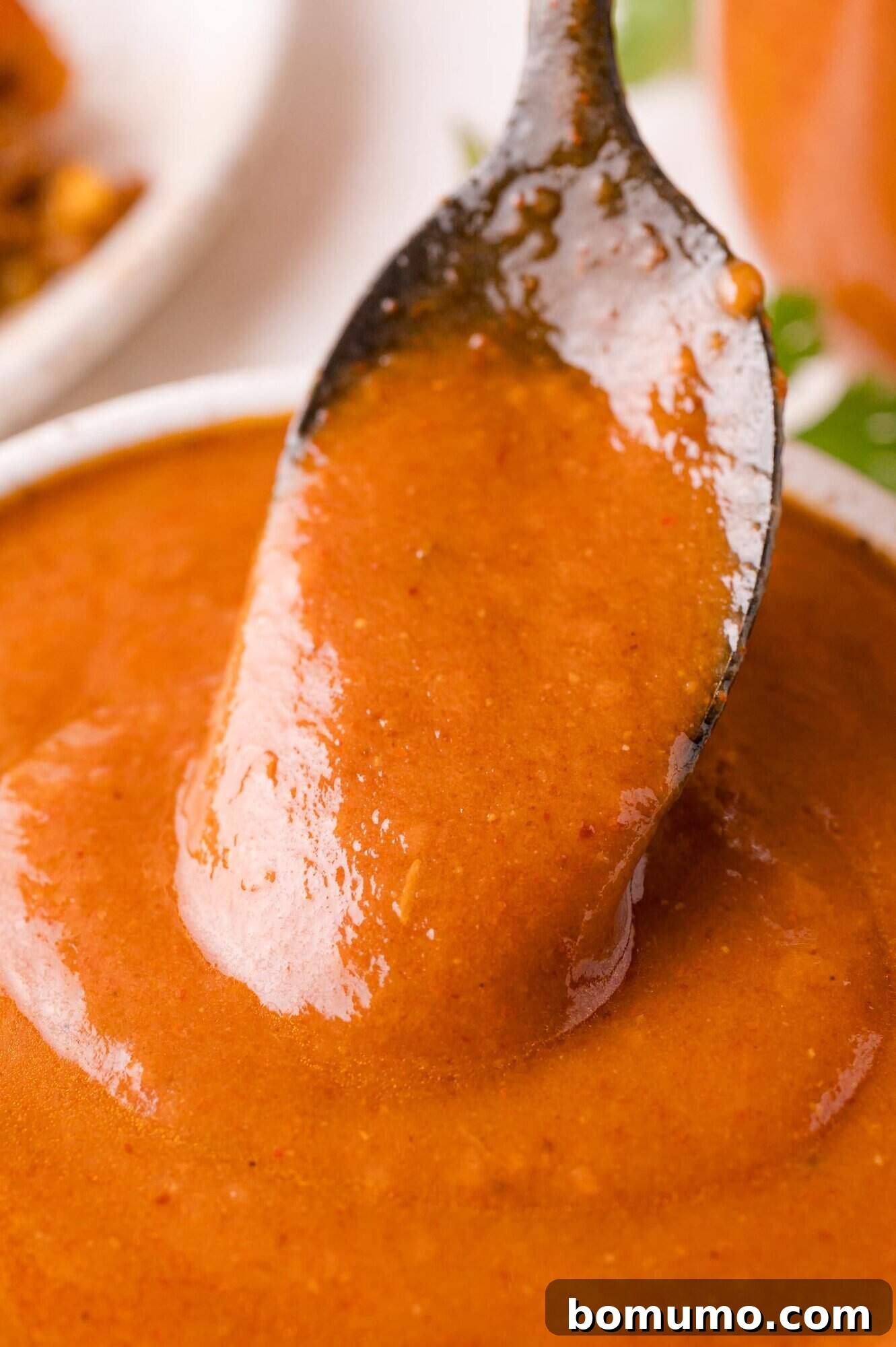 Close-up of a spoon lifting a stream of smooth, reddish-brown enchilada sauce from a white bowl, showcasing its rich texture.