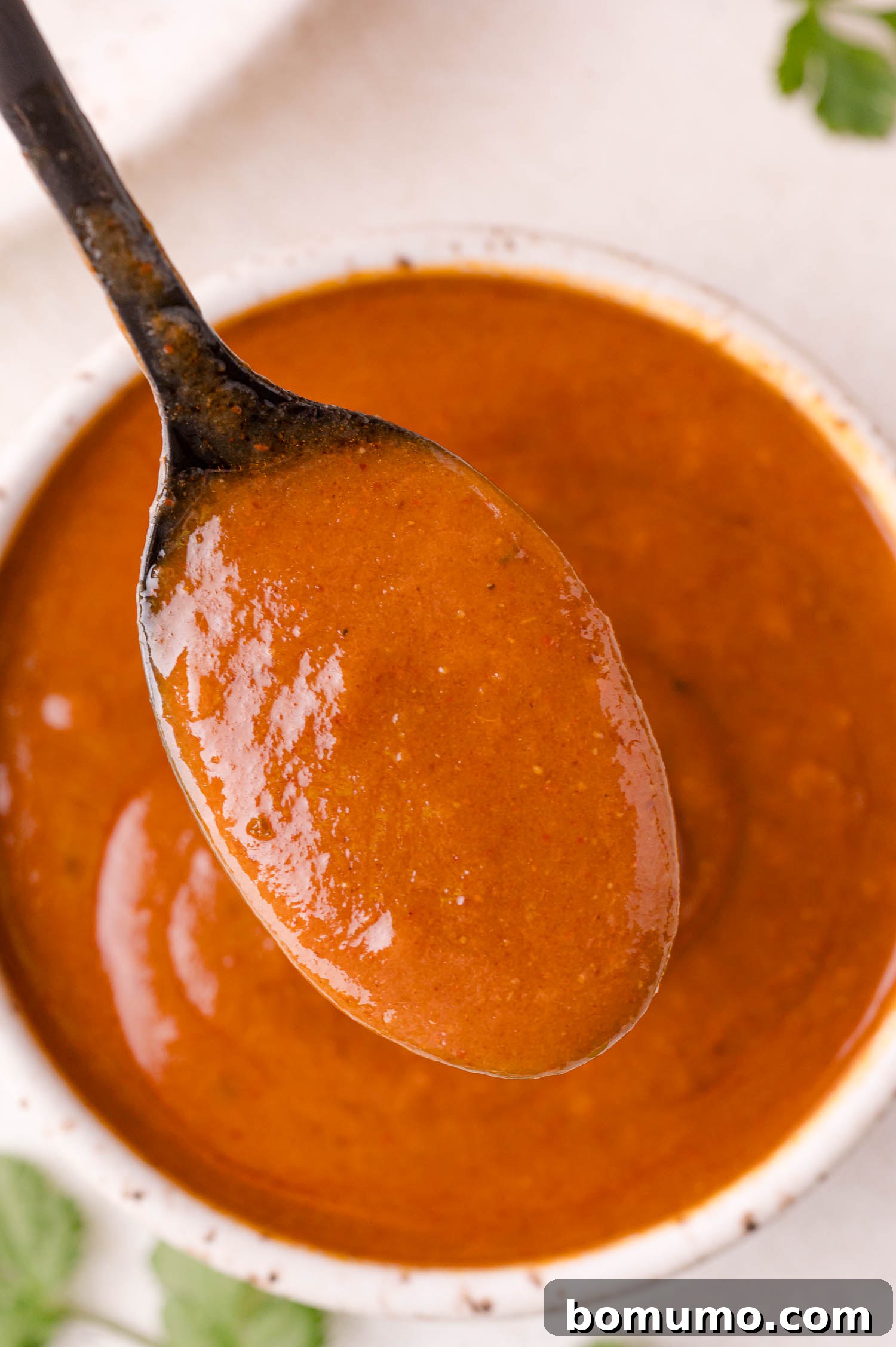 A spoon brimming with homemade enchilada sauce held over a white bowl filled with more of the rich, reddish-brown sauce.