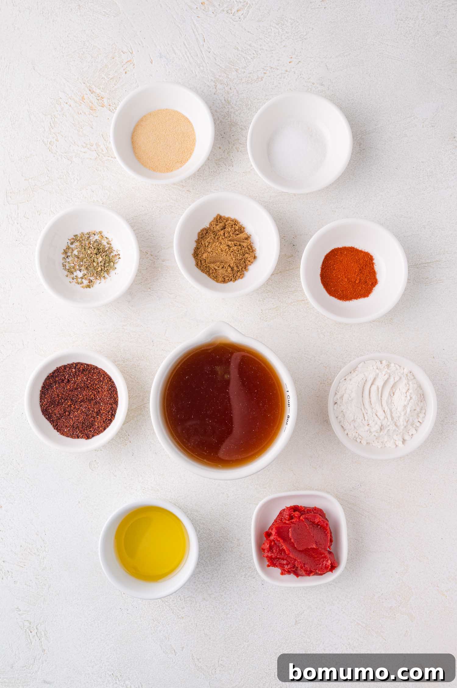 Various vibrant spices and essential ingredients, including chili powder, cumin, garlic powder, and tomato paste, are neatly arranged in small white bowls on a light-colored surface, ready for making enchilada sauce.