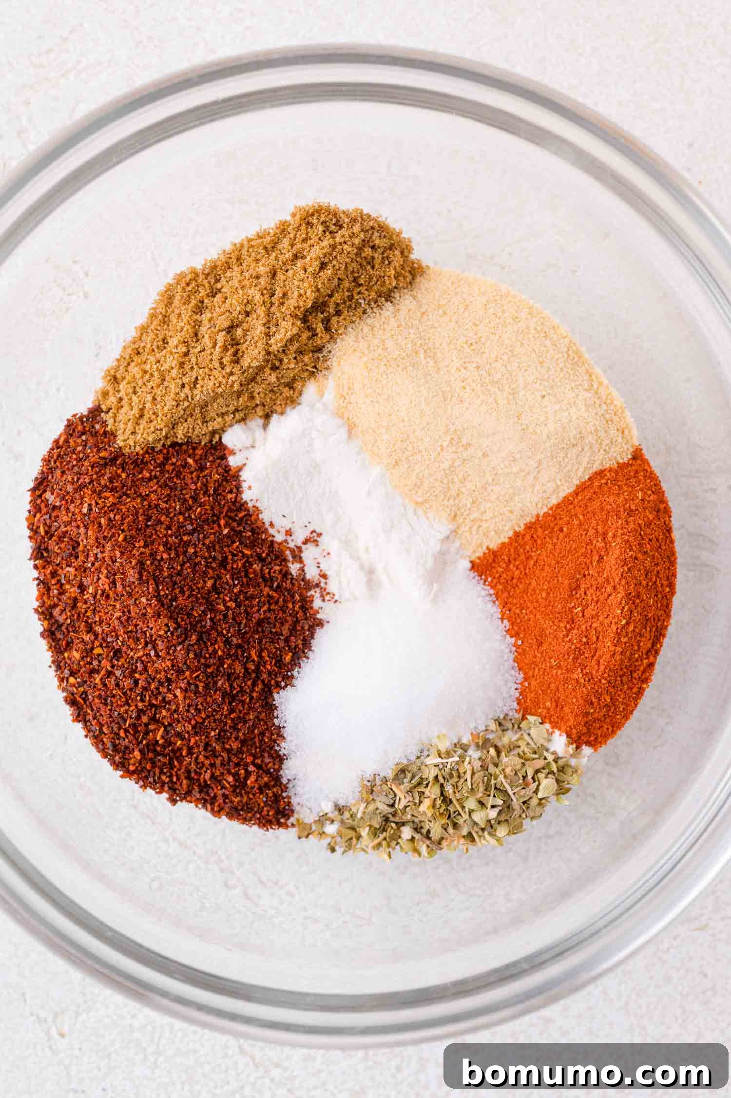 A glass bowl containing neatly arranged and separated spices including chili powder, cumin, garlic powder, onion powder, paprika, oregano, and salt, displayed on a light surface before mixing.