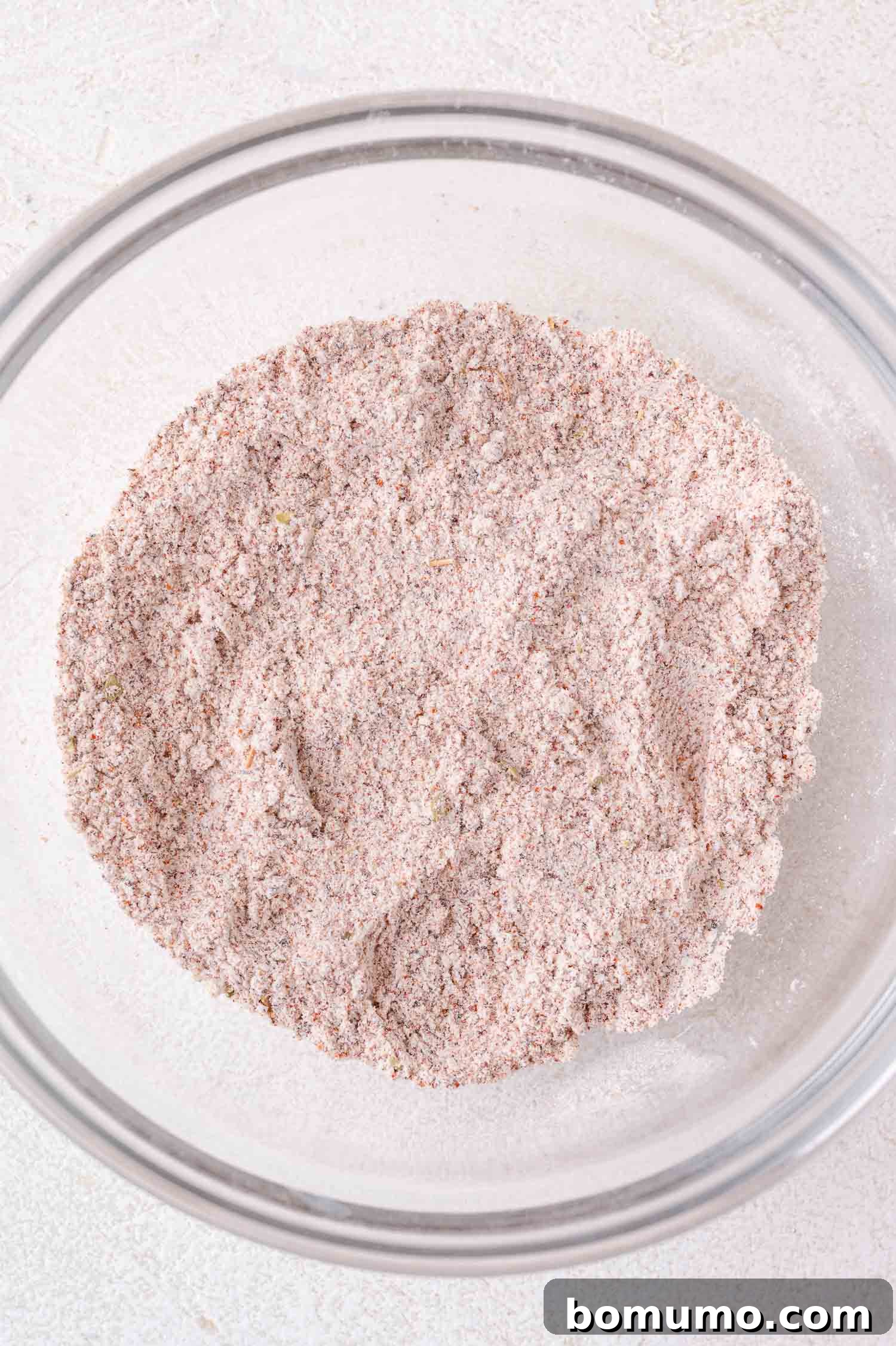 A glass bowl filled with a thoroughly mixed, dry, light brown blend of flour and various spices, showing a slightly uneven texture.