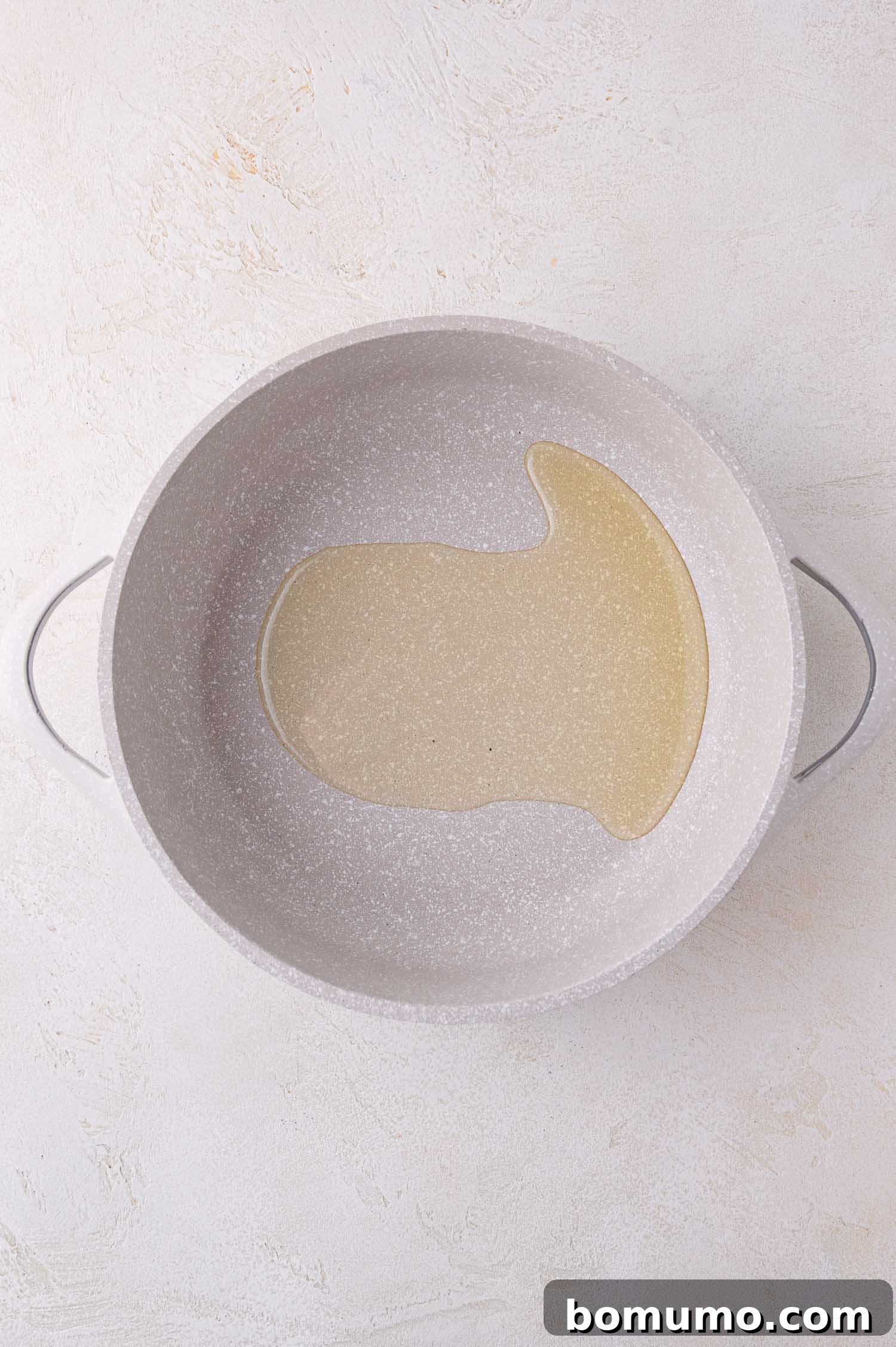 A large, white, speckled cooking pot viewed from above, with a clear pool of shimmering oil at its center, against a light textured background.