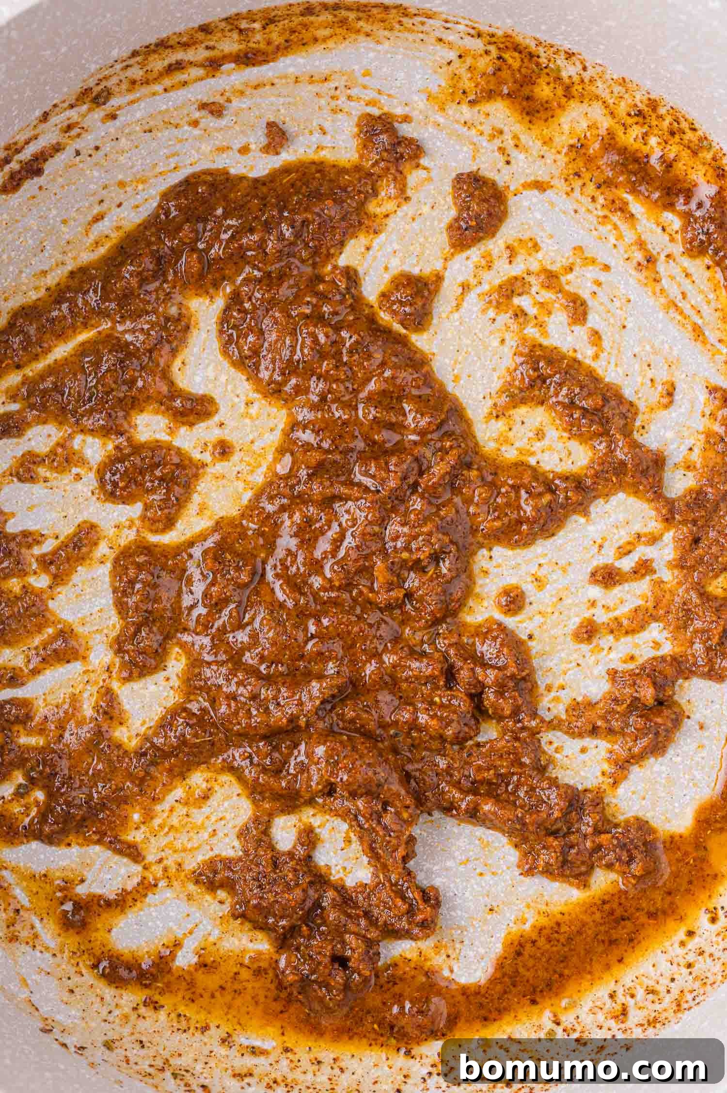 A thick, reddish-brown seasoning paste, a roux, is spread unevenly across the bottom of a light-colored pan, showing its rich texture.