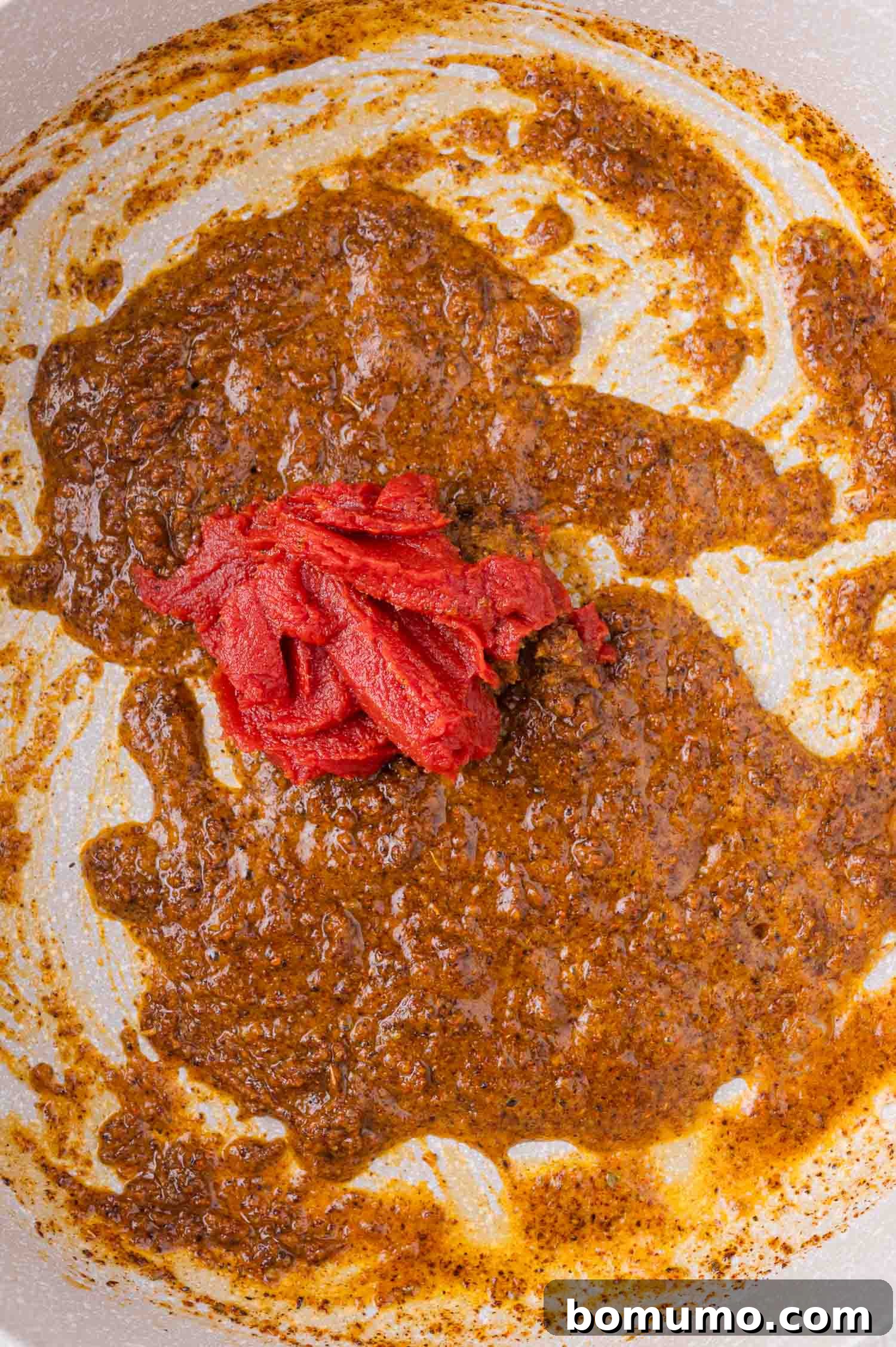 A dollop of vibrant red tomato paste rests on top of a brown roux mixture in a pan, ready to be combined.