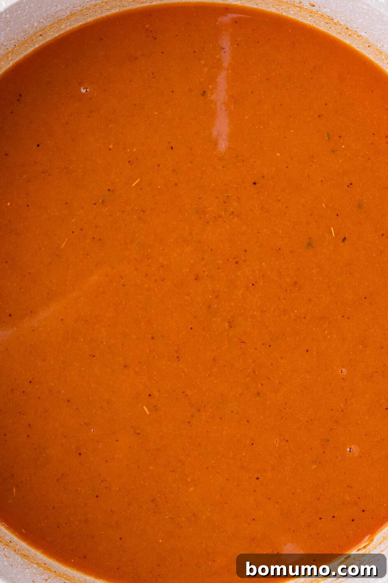 Close-up of a smooth, rich reddish-orange liquid enchilada sauce in a pan, showing visible flecks of seasoning and a slight sheen on its surface.