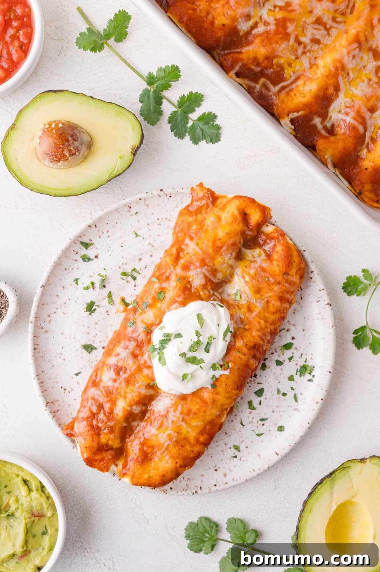 Hearty Beef Enchilada Bake 2 A plate with two enchiladas topped with sour cream and herbs, surrounded by avocado halves, cilantro, guacamole, and tomato salsa.