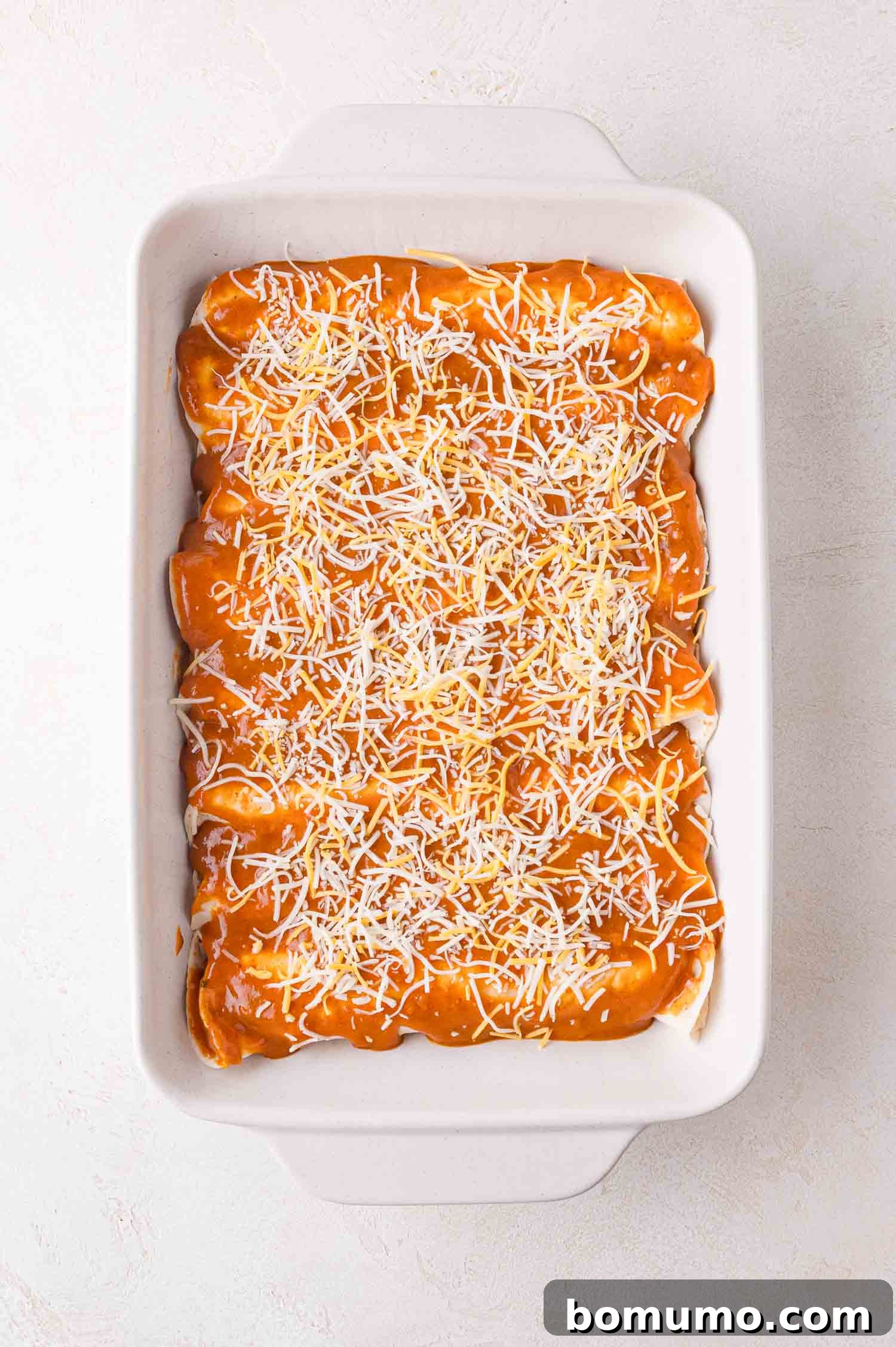 Hearty Beef Enchilada Bake 12 A white baking dish filled with enchiladas, topped with orange sauce and shredded cheese, set on a light textured surface.