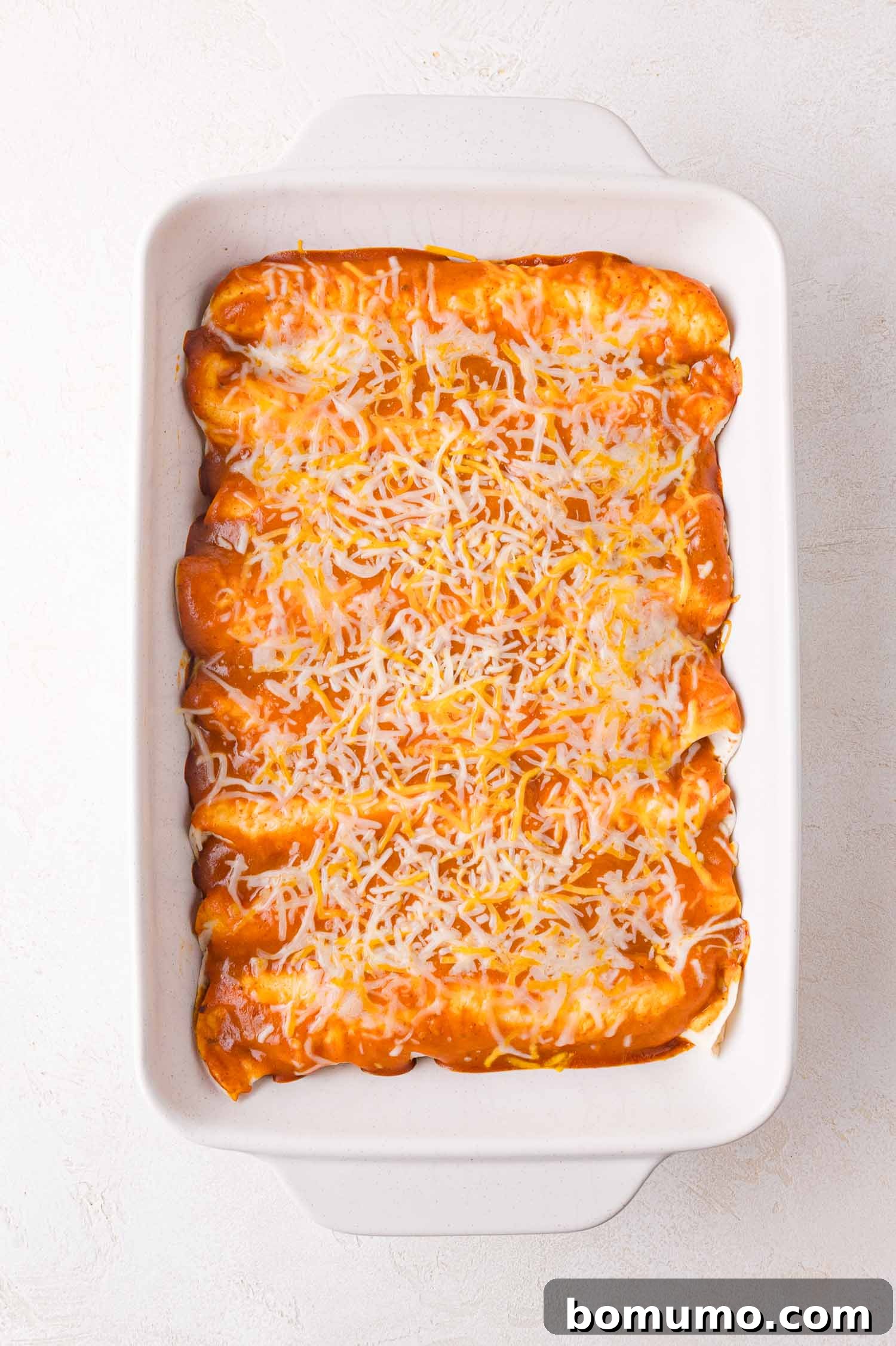 Hearty Beef Enchilada Bake 14 A white baking dish filled with enchiladas covered in red sauce and sprinkled with shredded cheese on a light background.