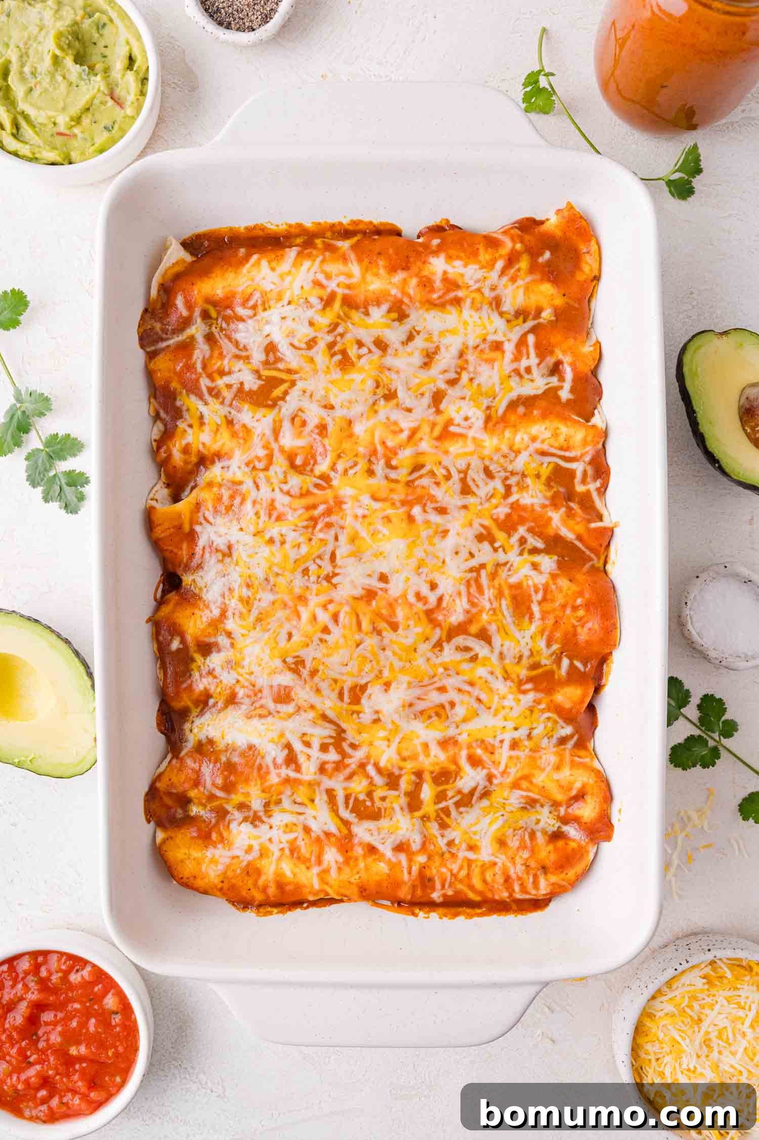 Hearty Beef Enchilada Bake 15 A casserole dish filled with red enchiladas topped with melted cheese. Surrounding the dish are avocado halves, cilantro, salsa, and guacamole.