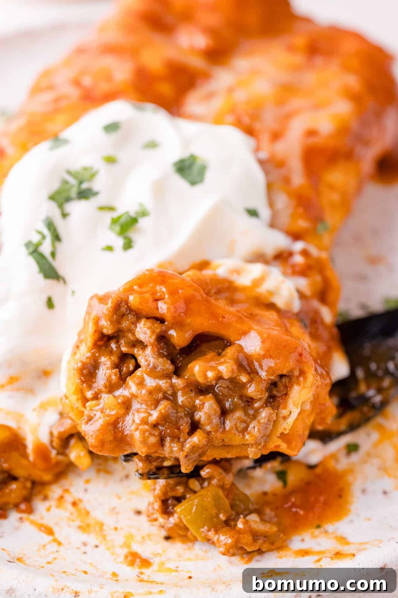 Hearty Beef Enchilada Bake 3 Close-up of a beef enchilada topped with melted cheese and sour cream, garnished with chopped herbs.