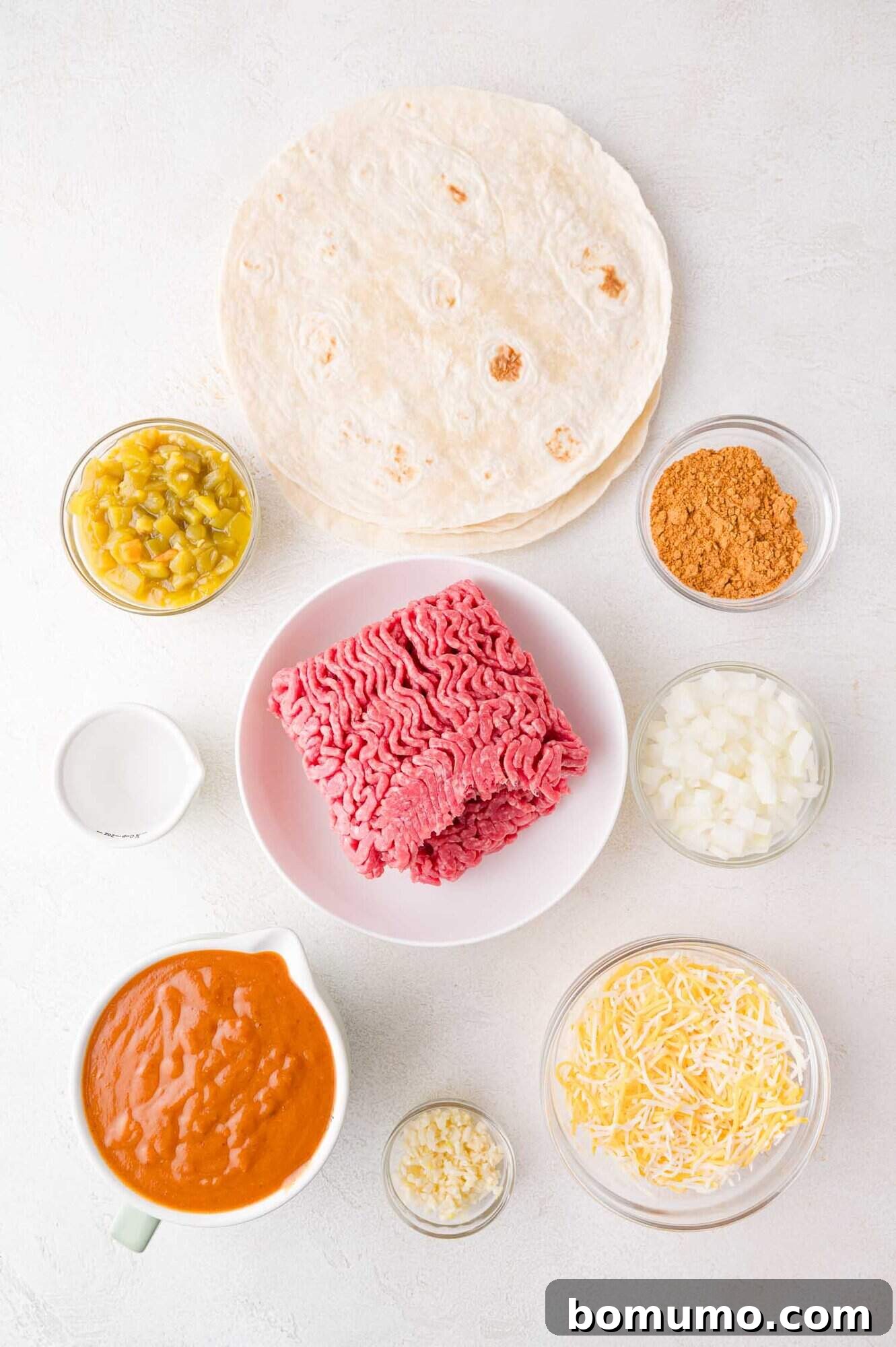 Hearty Beef Enchilada Bake 4 Ingredients for enchiladas include ground beef, tortillas, cheese, chopped onion, sauce, sour cream, refried beans, garlic, taco seasoning, and green chilies on a white surface.