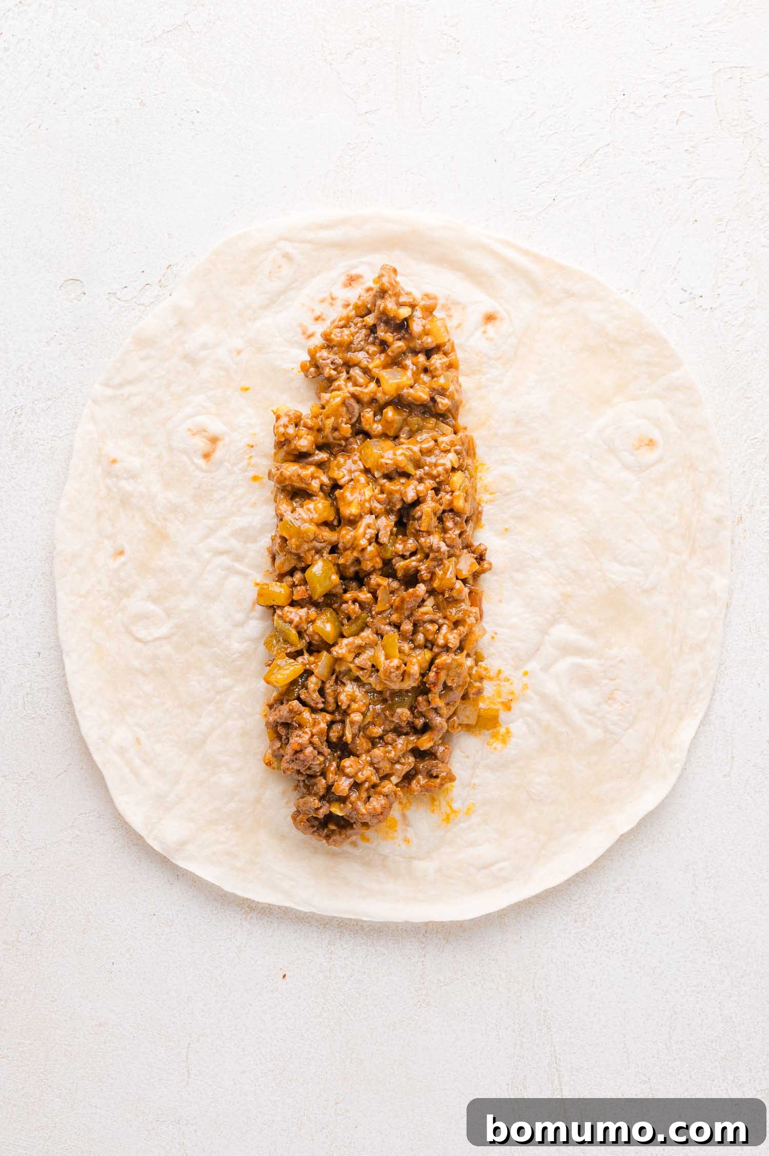 Hearty Beef Enchilada Bake 10 Uncooked enchilada on a light surface with a filling of ground meat, diced green peppers, and melted cheese placed in the center of a flour tortilla.