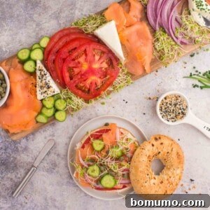 Bagel with smoked salmon, cucumber, tomato, onion, alfalfa sprouts, and cream cheese on a plate, next to a wooden board with more toppings, on a gray surface.