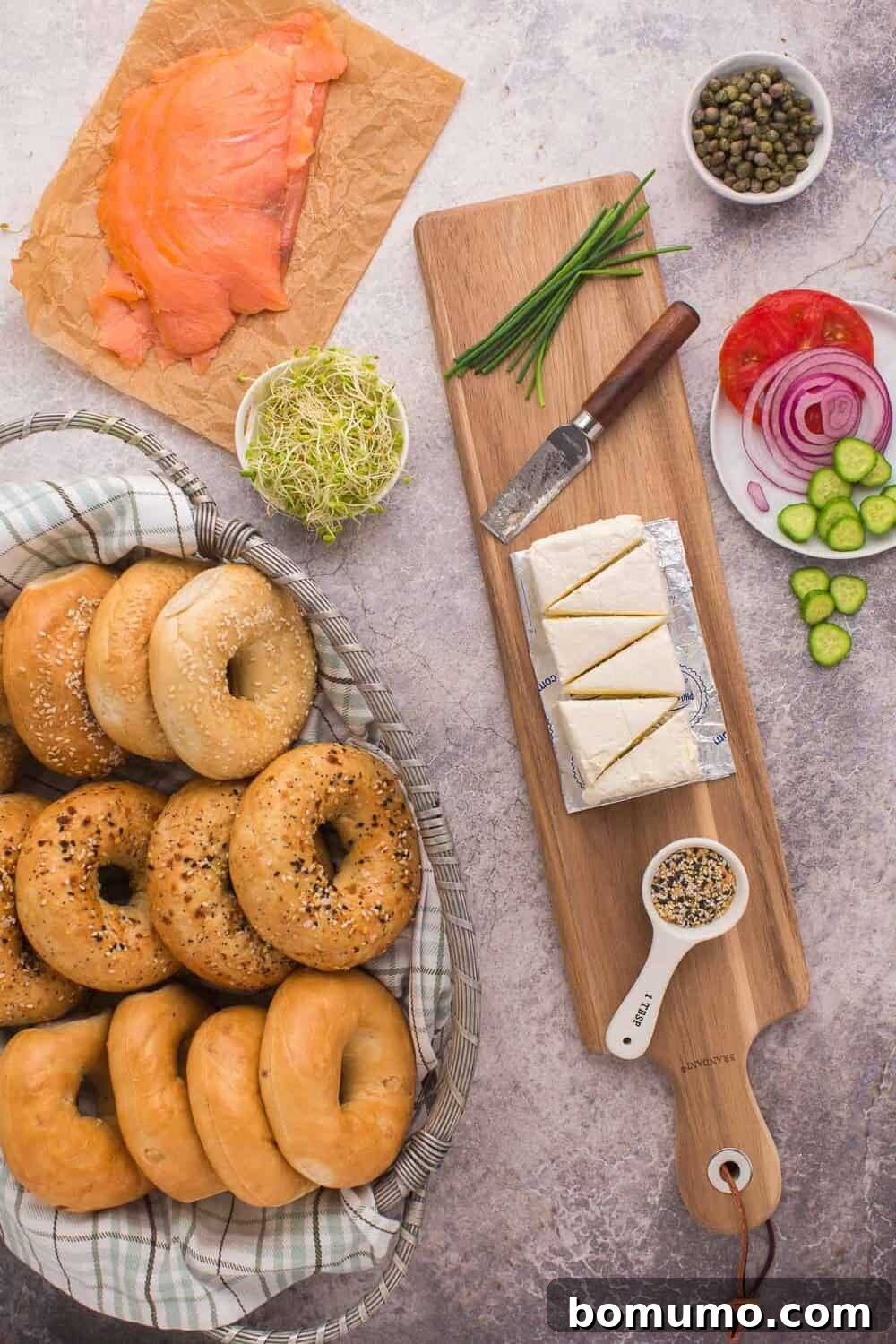 A comprehensive bagel and lox board arrangement. A basket of assorted bagels sits nearby. On a wooden board, there's a generous serving of smoked salmon, artfully cut cream cheese, fresh tomato slices, red onion, crisp cucumbers, capers, and alfalfa sprouts. A small knife and a sprinkle of chives add finishing touches, with everything bagel seasoning within reach.