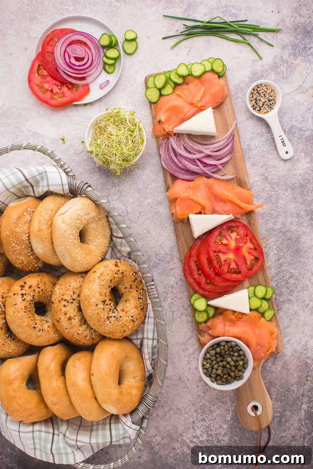 A delightful brunch tableau featuring a basket of freshly baked bagels beside a bountiful board. The board is adorned with rich smoked salmon, thinly sliced tomatoes, vibrant red onion, refreshing cucumber, creamy cheese, and delicate sprouts, all artfully garnished with various seeds and capers, ready for serving.
