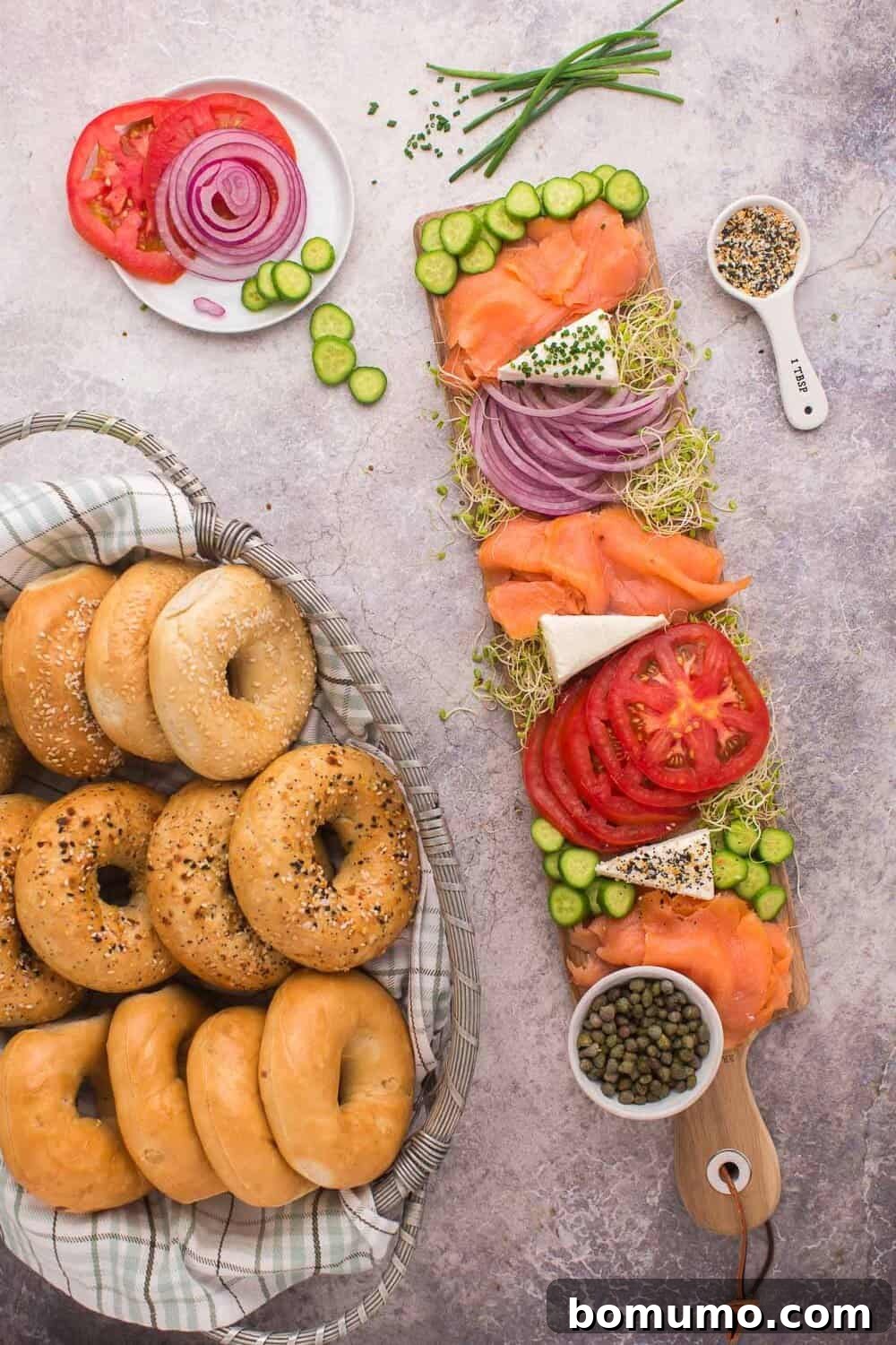 An abundant Bagel and Lox Board presentation. A basket filled with bagels rests beside a wooden board showcasing glistening smoked salmon, ruby-red tomato slices, thinly cut onions, crisp cucumber, and creamy cheese, accented with capers. A separate small plate offers additional tomato and onion slices for replenishment.