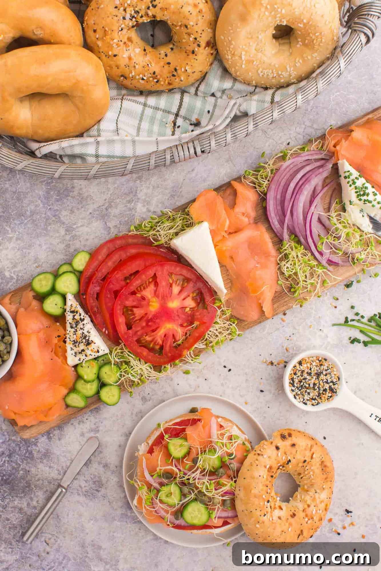 A delectable platter featuring assembled bagels with smoked salmon, fresh tomato slices, crisp cucumbers, thinly sliced red onions, creamy cream cheese, and delicate sprouts. Alongside, a rustic basket holds more bagels, and a small bowl offers seasoning for customization, all presented on a elegant gray surface.
