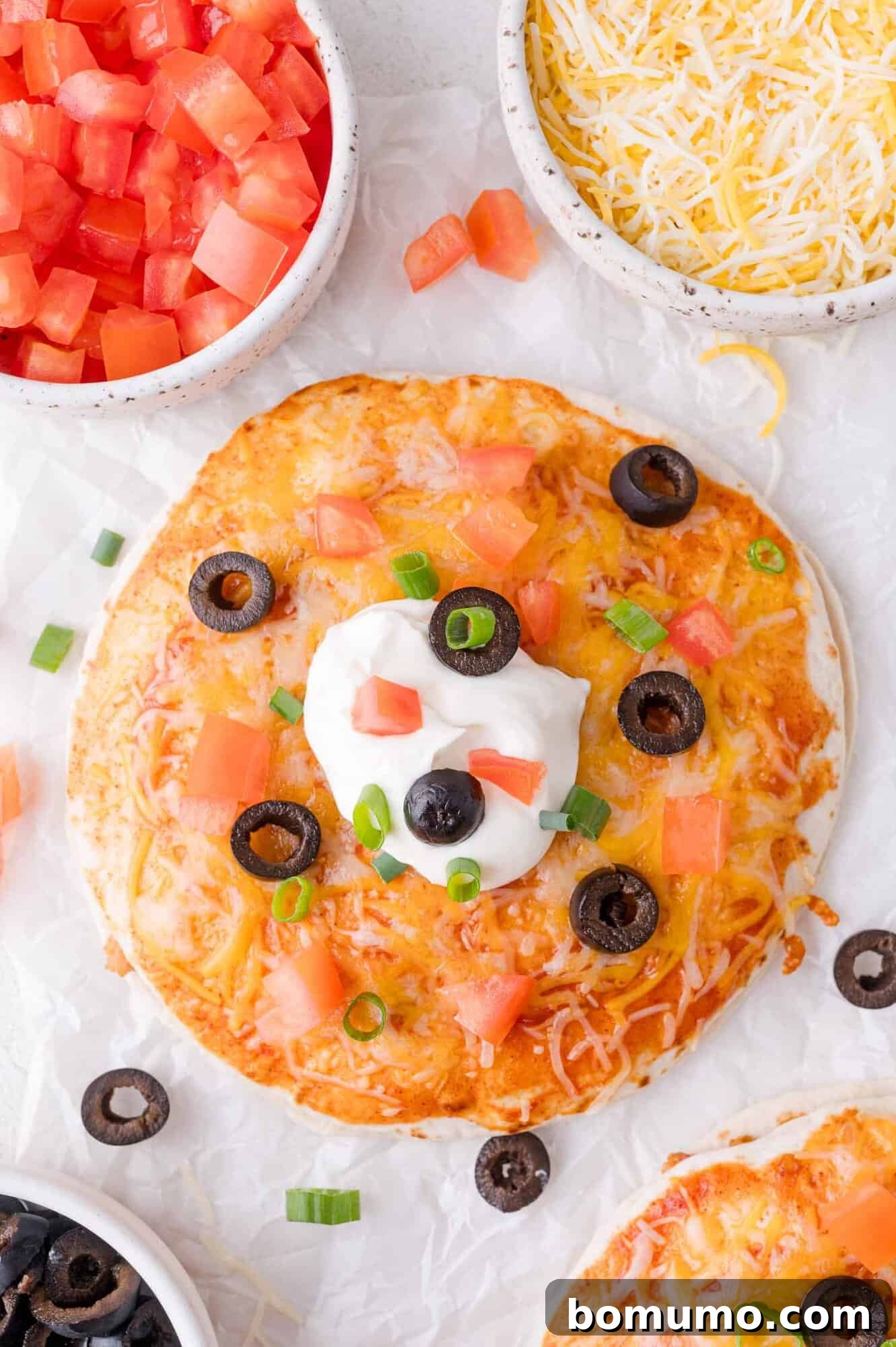 Crispy flour tortilla topped with melted cheese, diced tomatoes, a dollop of sour cream, black olives, and vibrant green onions, surrounded by bowls of fresh ingredients on parchment paper, ready to serve.