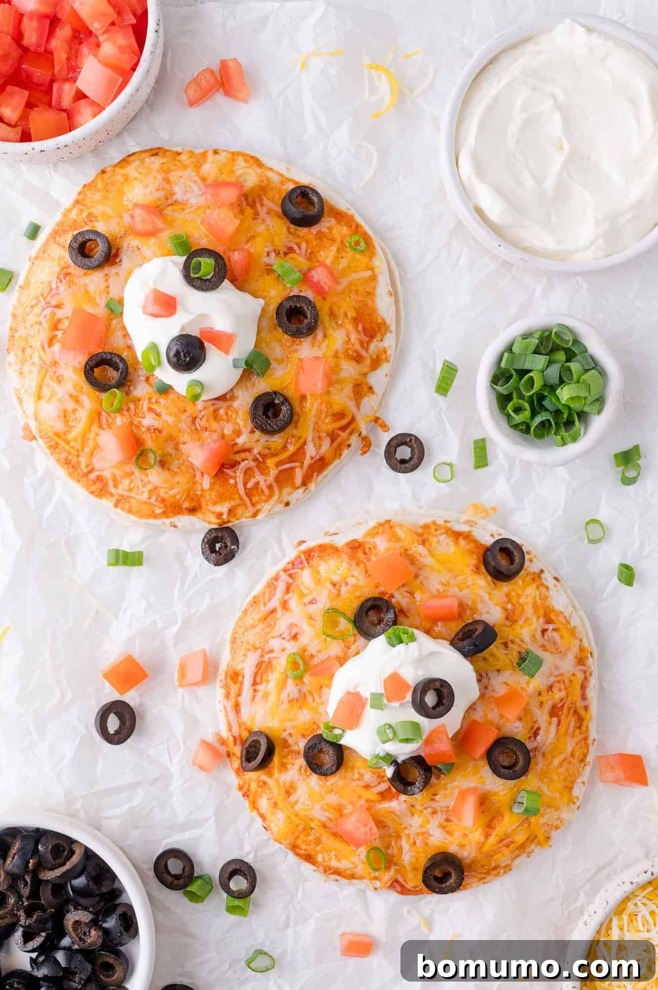 Two freshly baked tortilla pizzas, beautifully topped with melted cheese, diced tomatoes, black olives, sour cream, and green onions, presented on a white textured surface with accompanying small bowls of fresh toppings nearby.