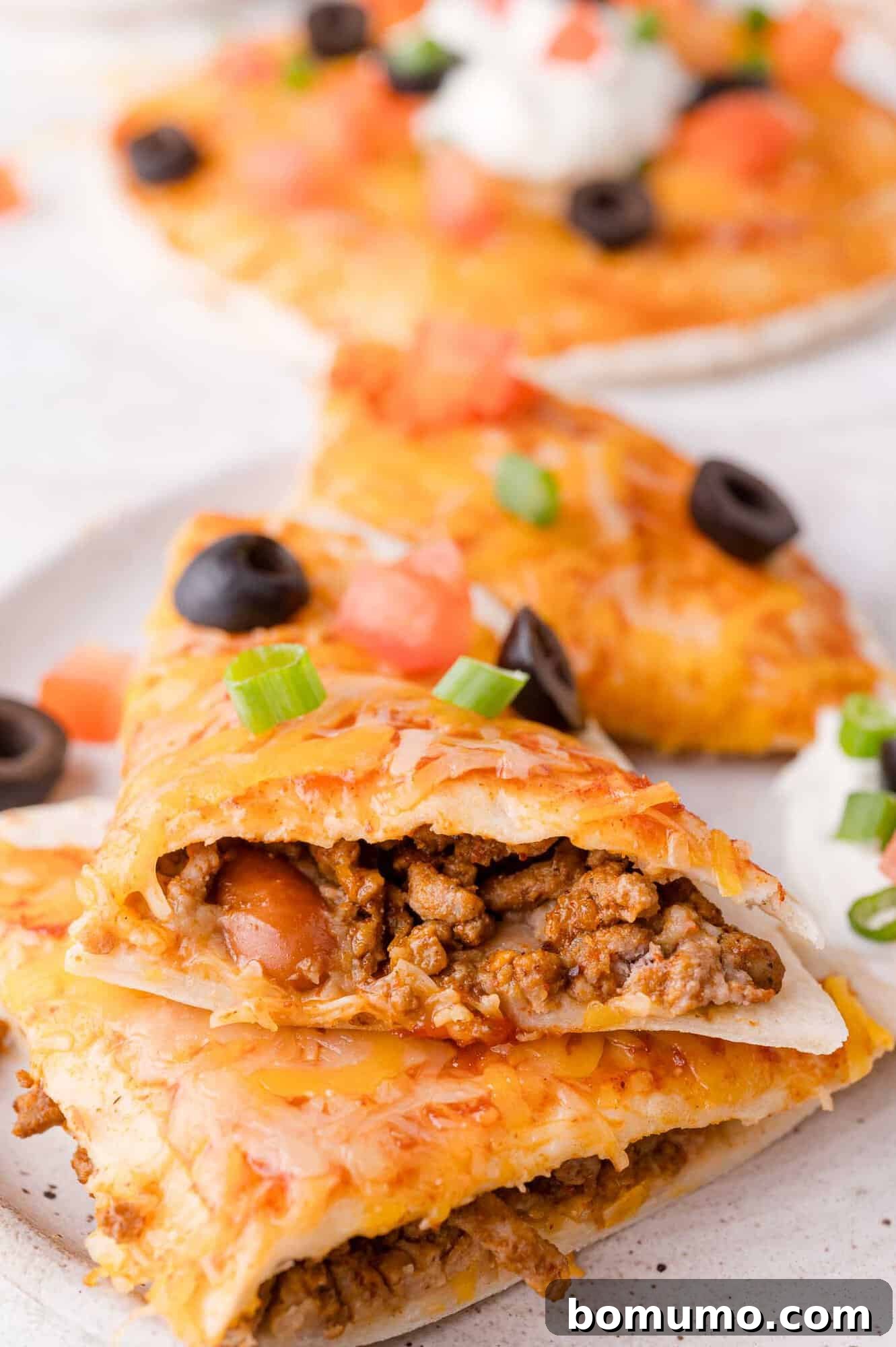 A close-up shot of a perfectly cut slice of Mexican pizza, revealing layers of savory ground beef, melted cheese, black olives, diced tomatoes, a dollop of sour cream, and fresh green onions. Other slices are visible in the soft-focus background.