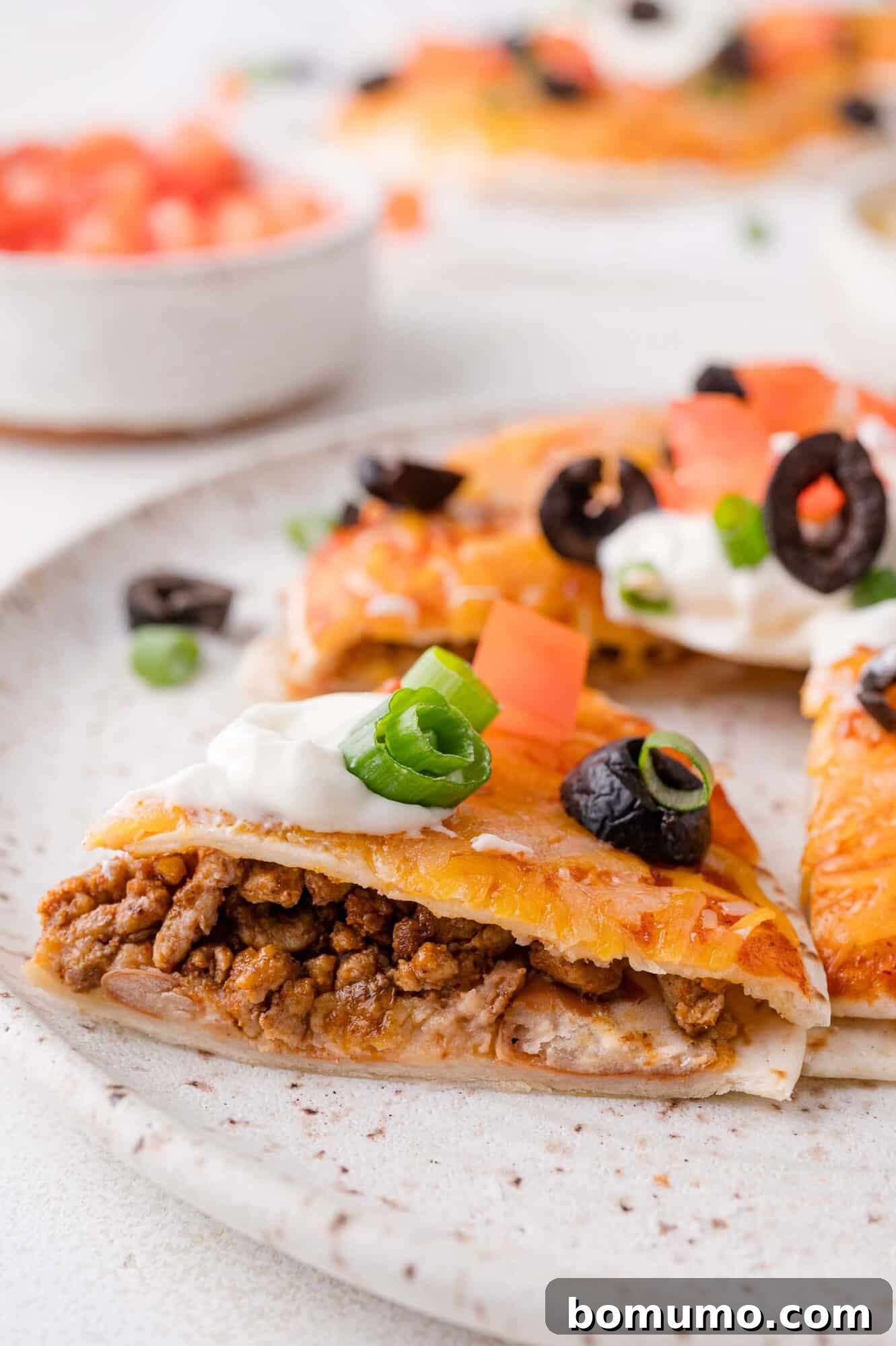 Close-up view of a perfectly baked beef Mexican pizza slice, garnished with a dollop of creamy sour cream, vibrant diced tomatoes, thinly sliced black olives, and fresh green onions, resting invitingly on a white plate with other slices in the background.