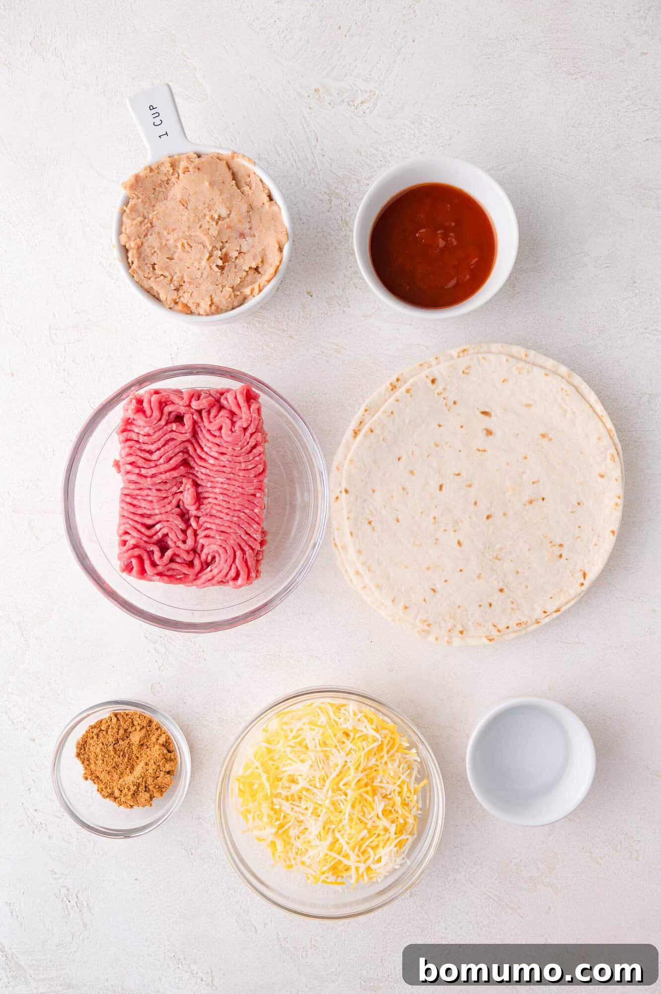 A meticulous arrangement of fresh ingredients on a clean white surface: raw ground meat, soft tortillas, shredded Mexican blend cheese, creamy refried beans, vibrant salsa, taco seasoning, and an empty measuring cup, all ready for the culinary process.