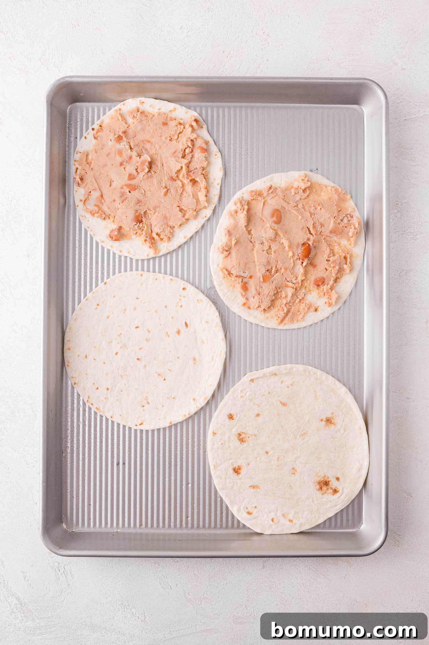 Four golden flour tortillas neatly arranged on a metal baking sheet. Two are generously spread with mashed refried beans, while the other two await their delicious topping.