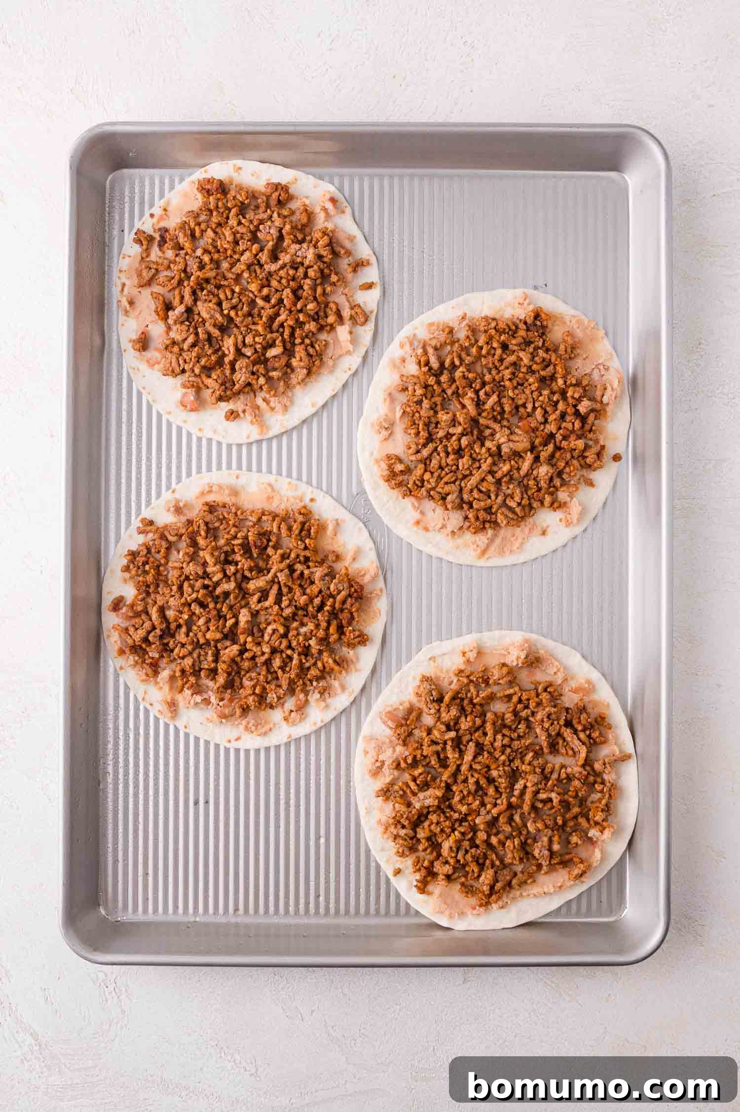 Four round tortilla bases, each crowned with a savory layer of perfectly spiced ground meat, are neatly arranged on a metal baking tray, ready for the next delicious step.