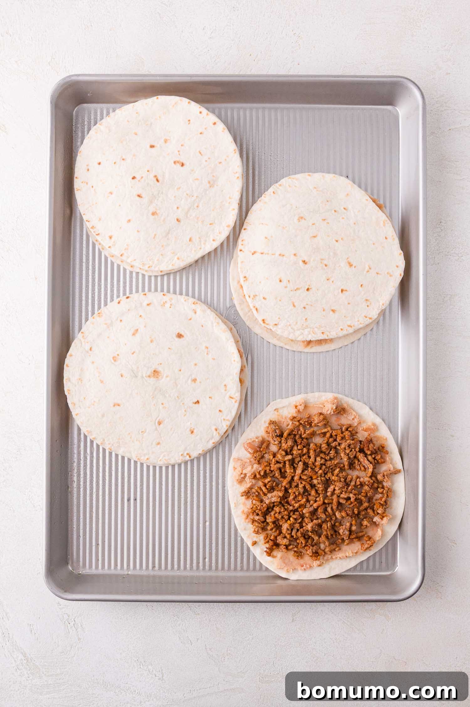 Four tortilla stacks are arranged on a baking sheet. Three are now topped with a second plain tortilla, while one in the foreground still reveals the cooked ground meat layer, illustrating the assembly process.
