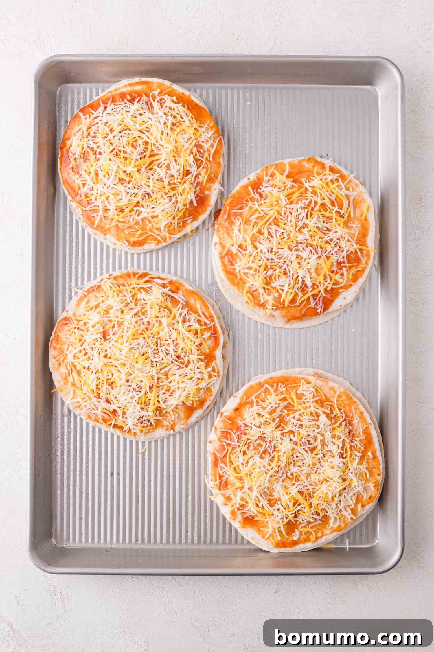 Four individual, unbaked Mexican pizzas, each generously topped with a thin layer of vibrant tomato sauce and a sprinkle of shredded cheese, are perfectly aligned on a baking tray, ready to go into the oven.