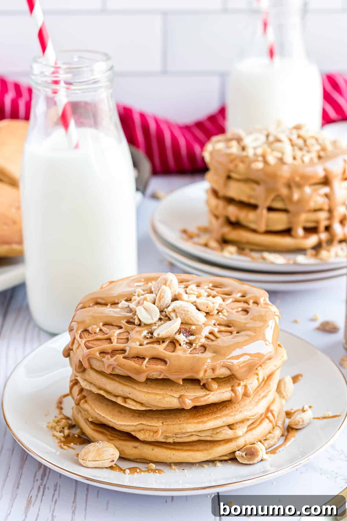 A glorious stack of fluffy peanut butter pancakes, generously drizzled with melted peanut butter and adorned with crunchy chopped peanuts, resting on a pristine white plate. In the blurred background, a bottle of milk and a cheerful red straw hint at a complete breakfast experience.